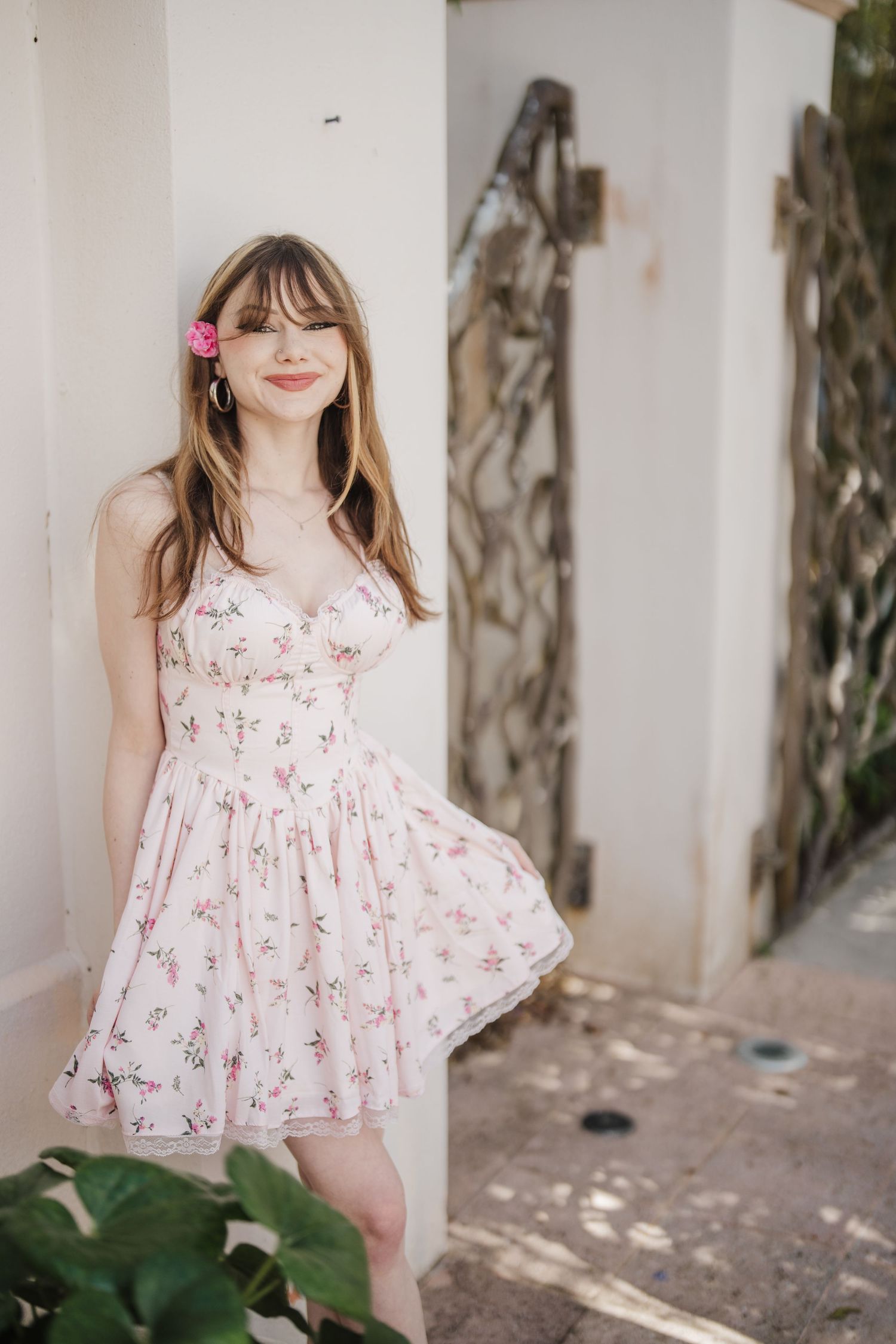 A person wearing a delicate white and pink floral print summer dress poses against a light-colored wall with greenery.