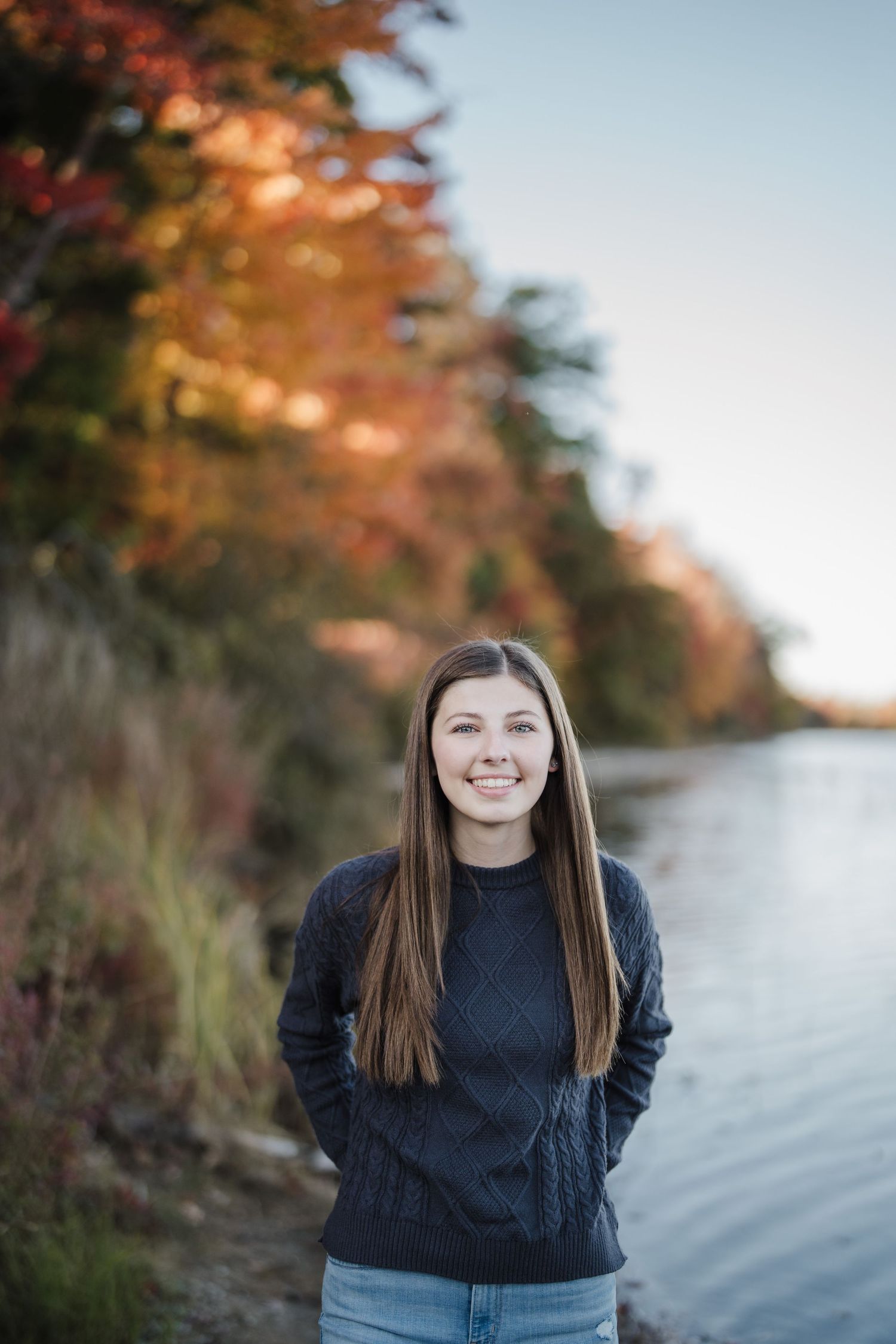 A person in a navy cable knit sweater stands by a lake with autumn foliage in the background during golden hour.