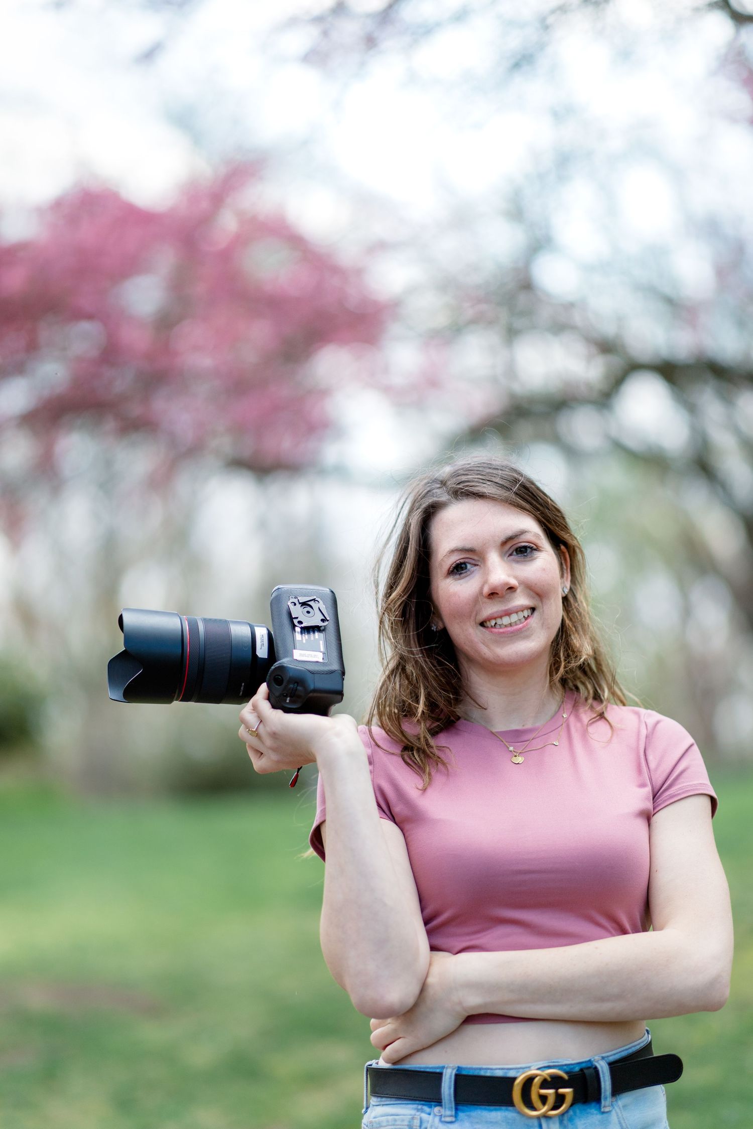 Woman photographer smiling outdoors holding DSLR camera, wearing pink top and Gucci belt, with blooming trees background.