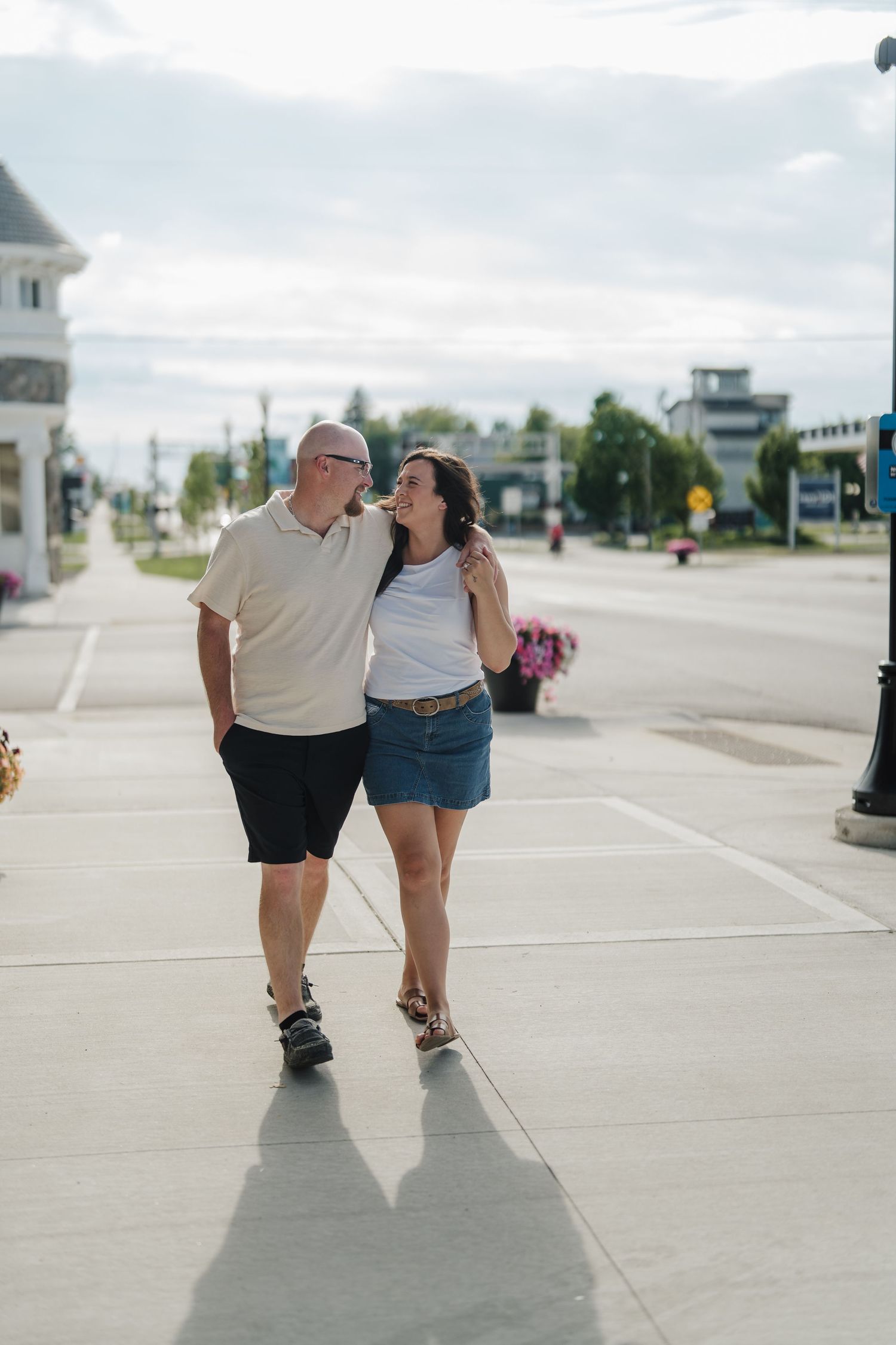 A couple walks arm in arm on a sunny sidewalk, smiling at each other in a small town setting.