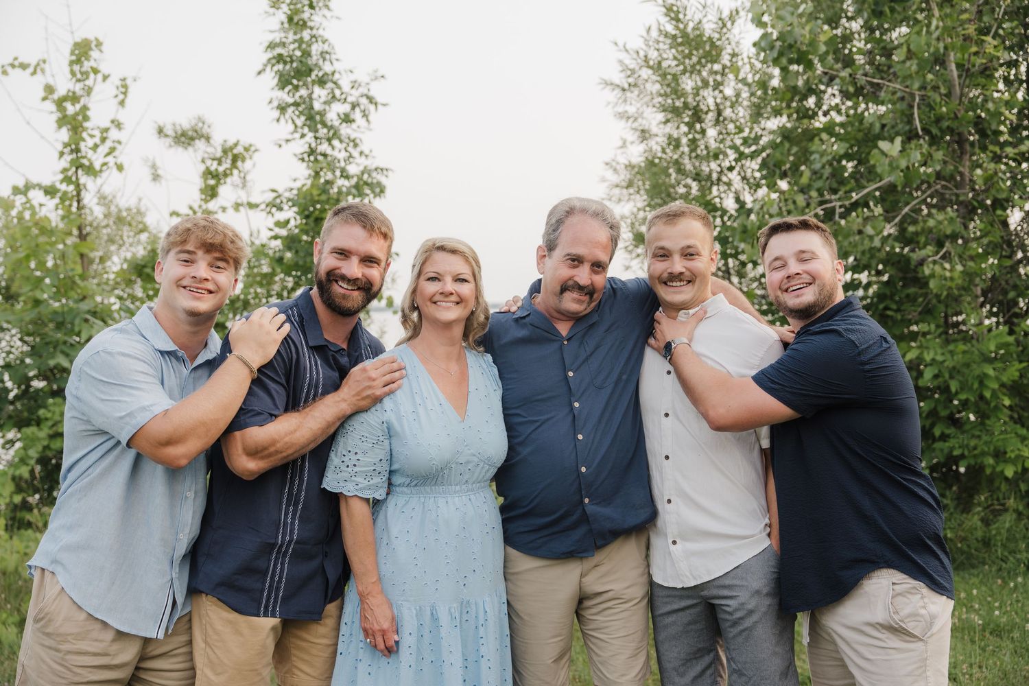 A smiling family of six poses outdoors among green trees, wearing casual blue and neutral outfits in a warm portrait.