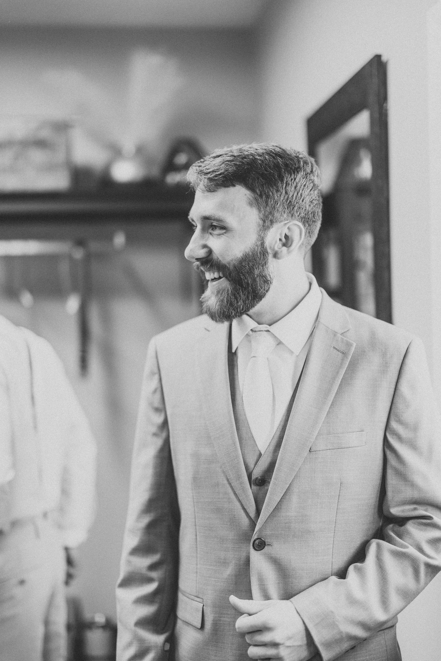 Black and white portrait of groom in grey three-piece suit smiling indoors.