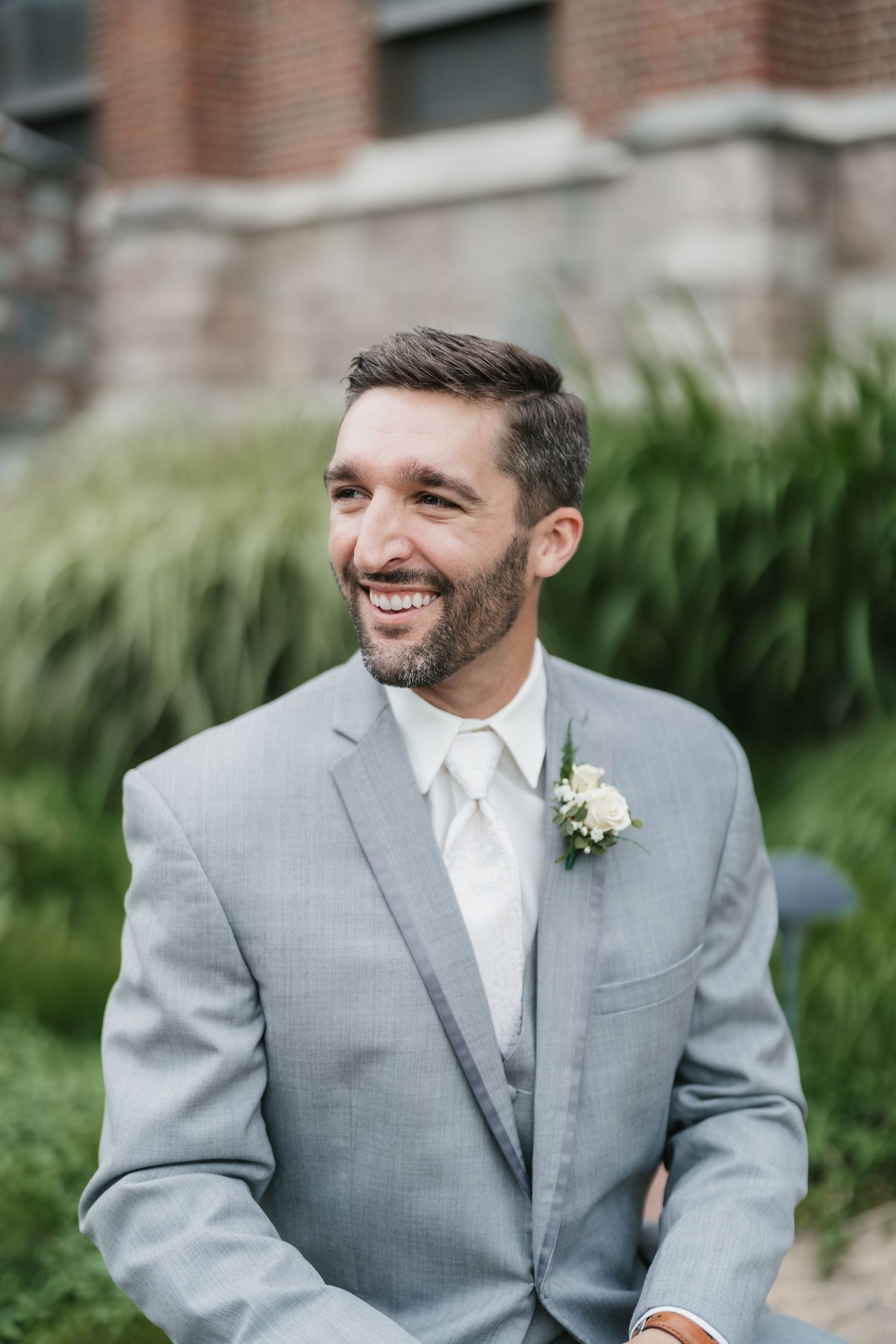 Groom in light grey suit with white boutonniere smiling outdoors.