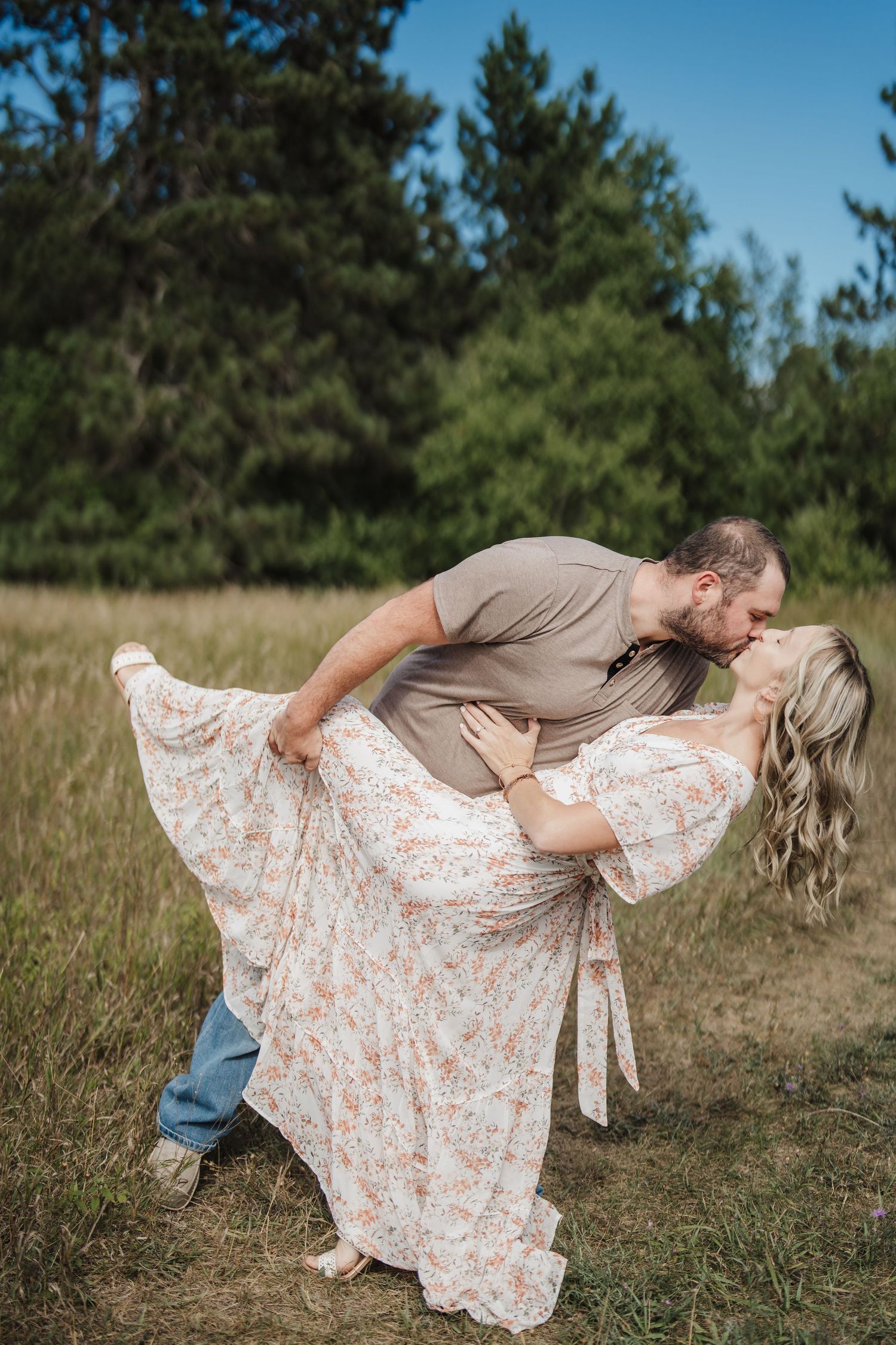 A romantic couple in a field shares a tender dip pose, with her floral dress flowing gracefully in the summer setting.