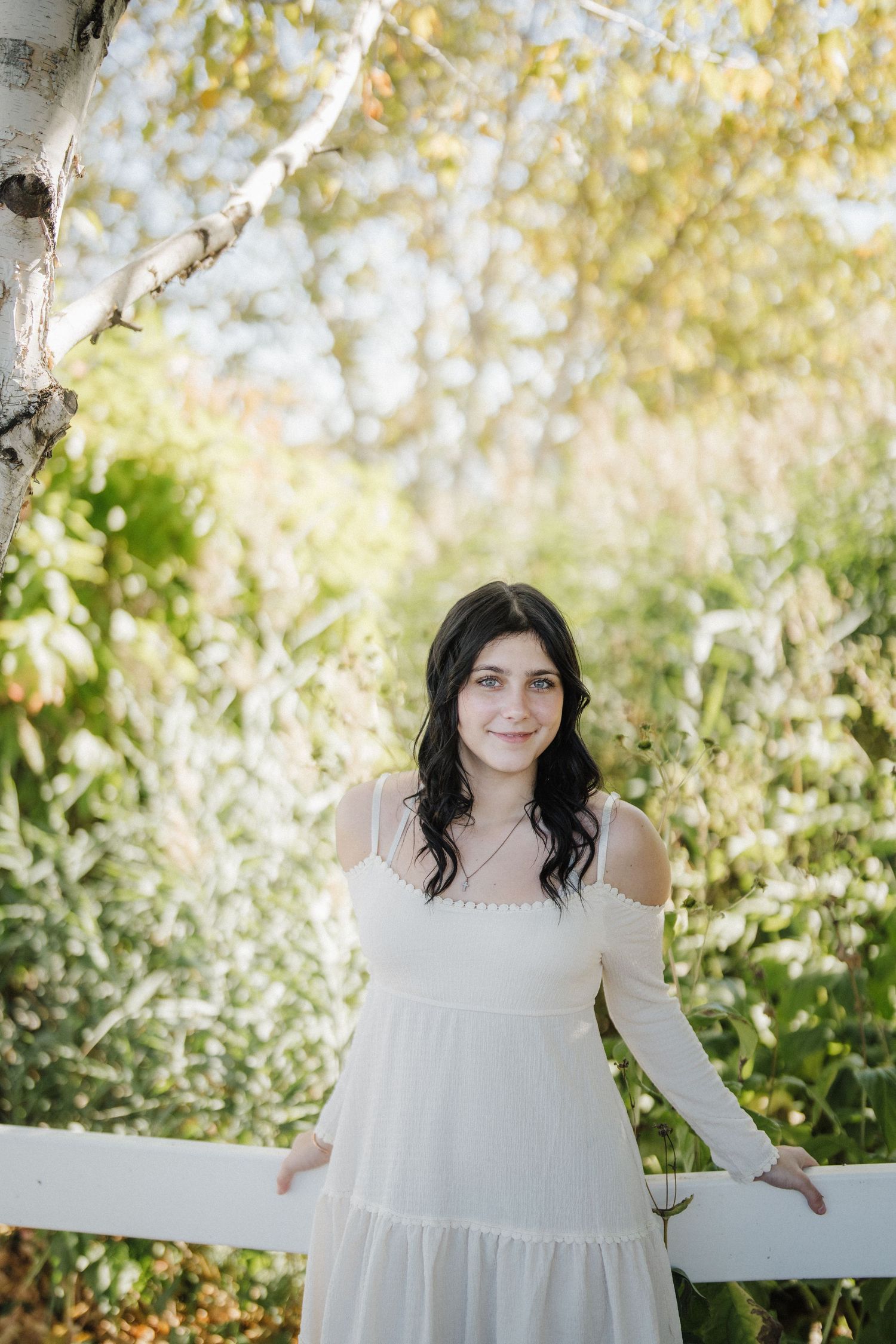 A summer portrait by a white fence with someone wearing a flowing white dress against birch trees.