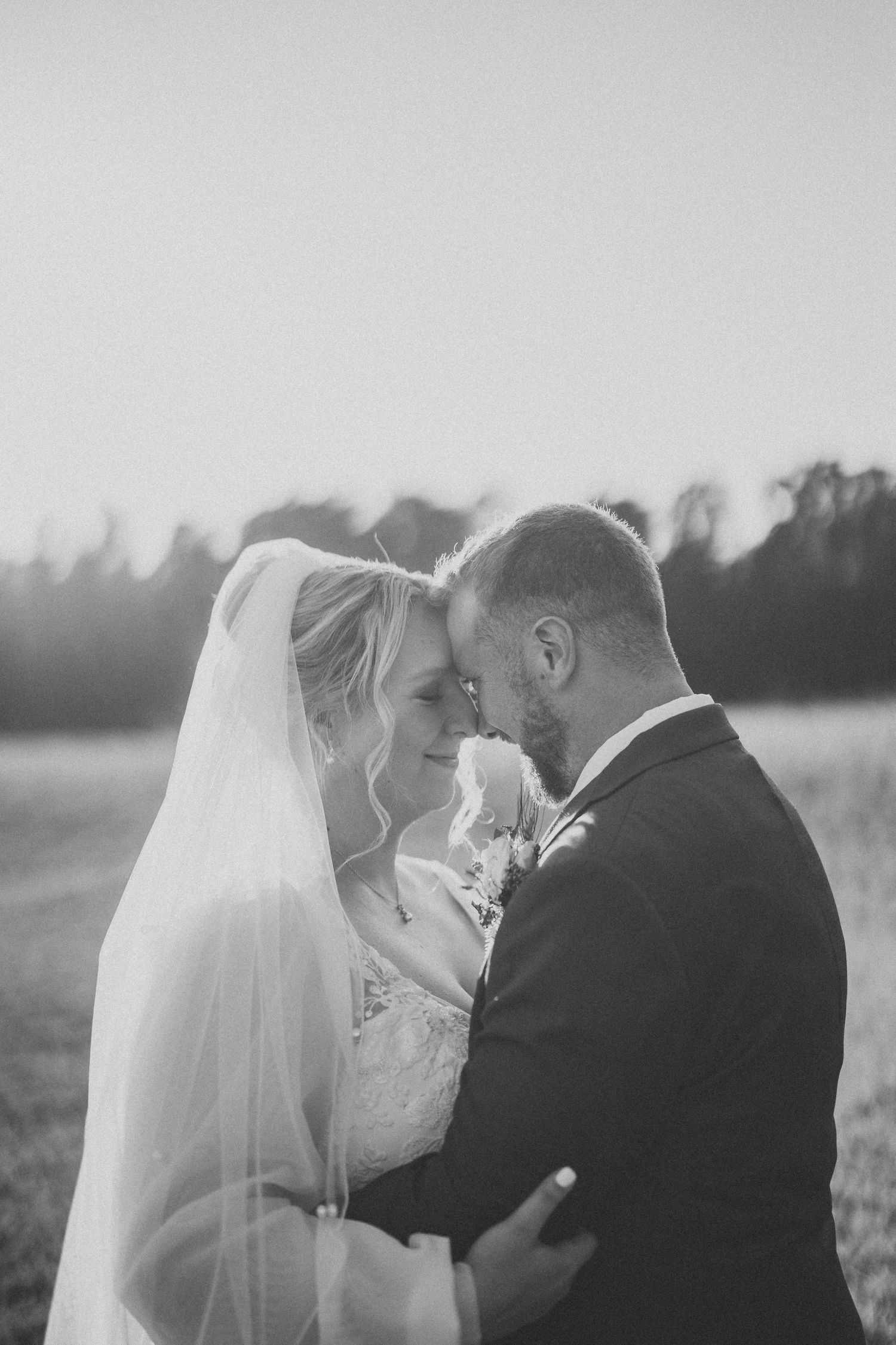 A romantic wedding couple wearing protective face masks share a tender moment in a field at sunset.