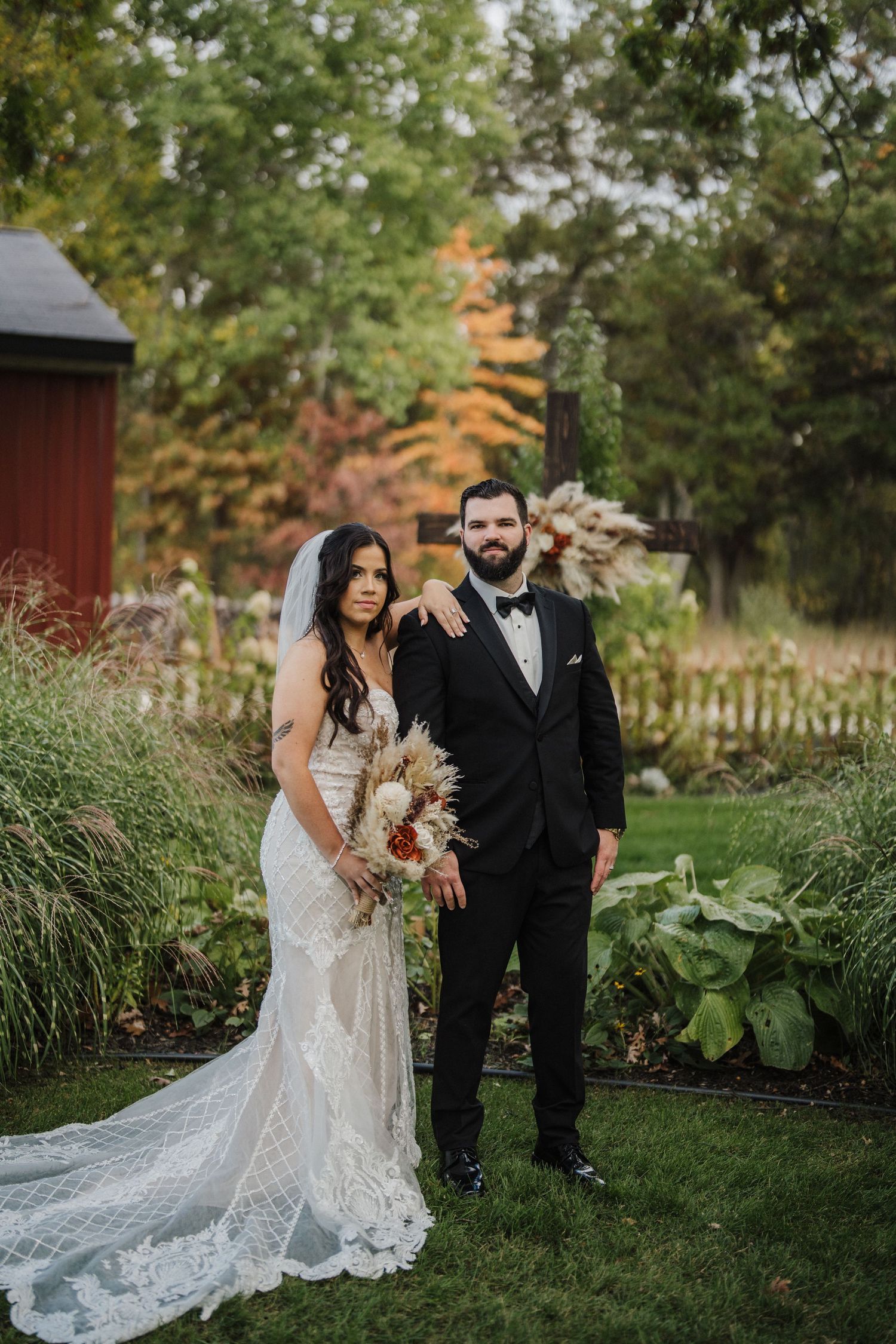 A bride in a lace gown with long train poses with her partner in a garden setting with rustic red buildings.