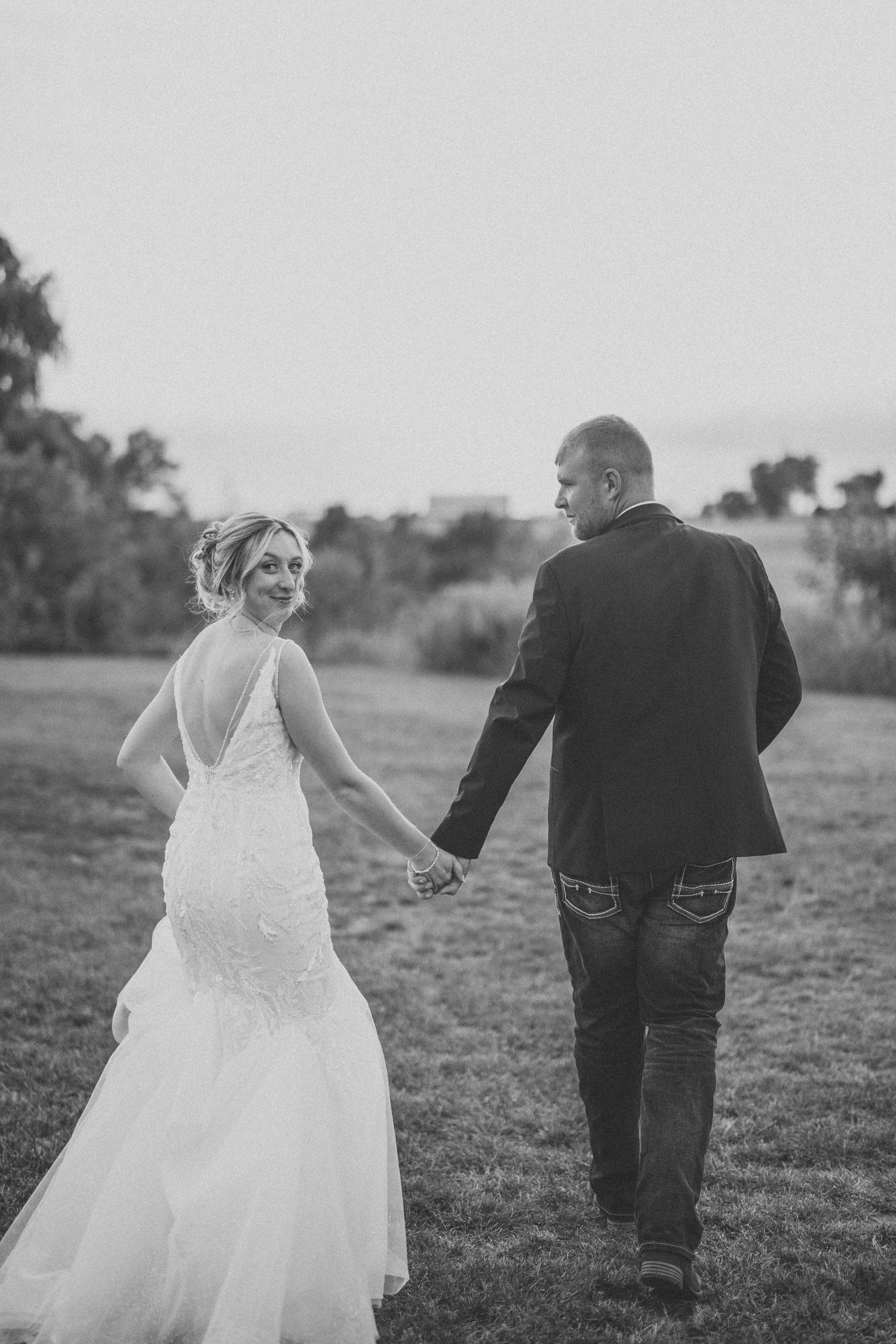 Black and white wedding photo of couple holding hands while walking together in an outdoor field at sunset.