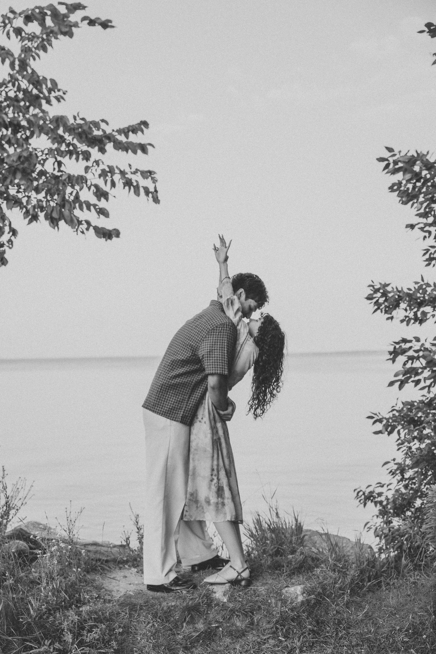 Black and white photo of a couple kissing by a lakeside, surrounded by trees, as the woman raises her hand.