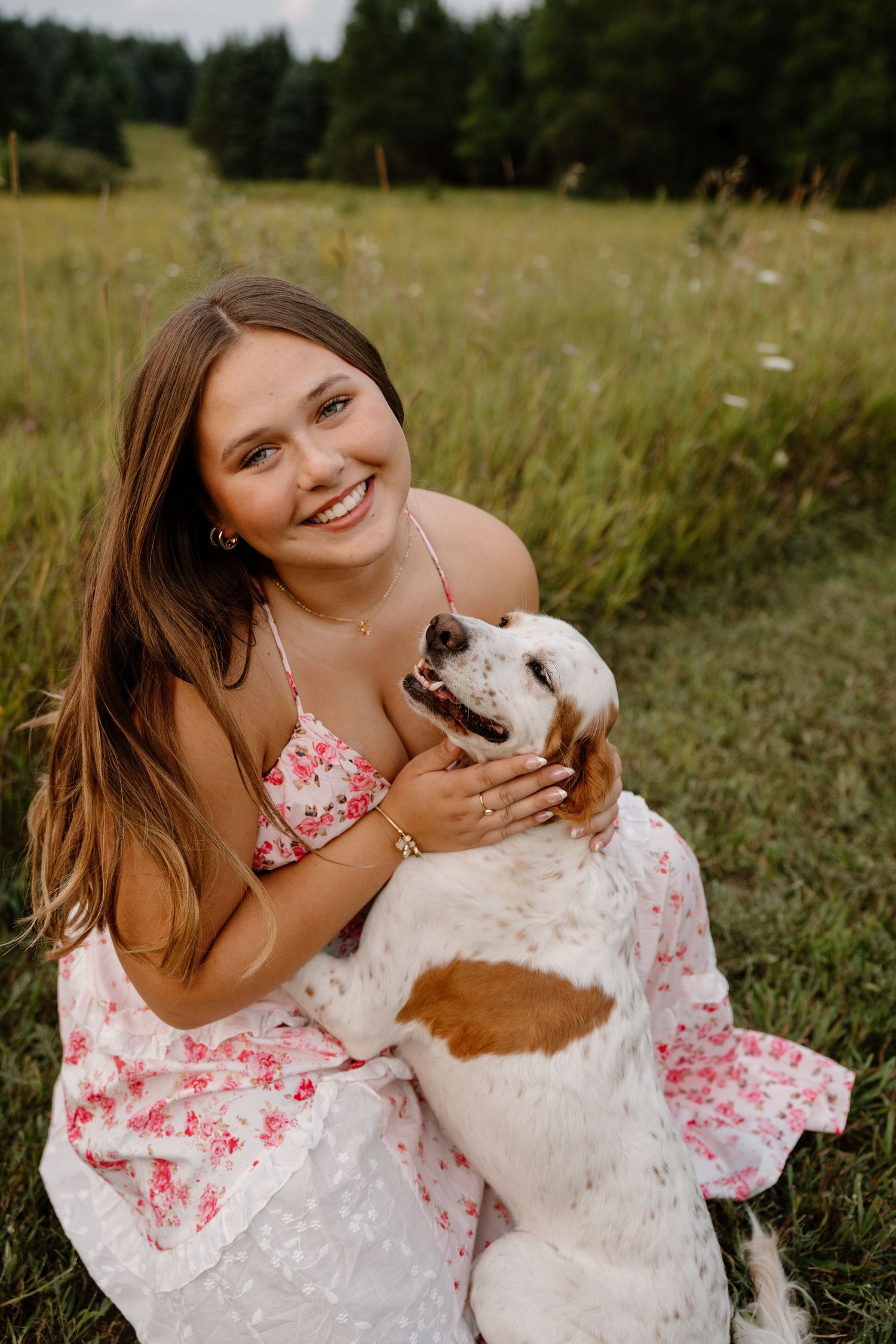 Smiling young woman in floral dress sitting outdoors in a meadow, hugging her brown and white beagle dog.