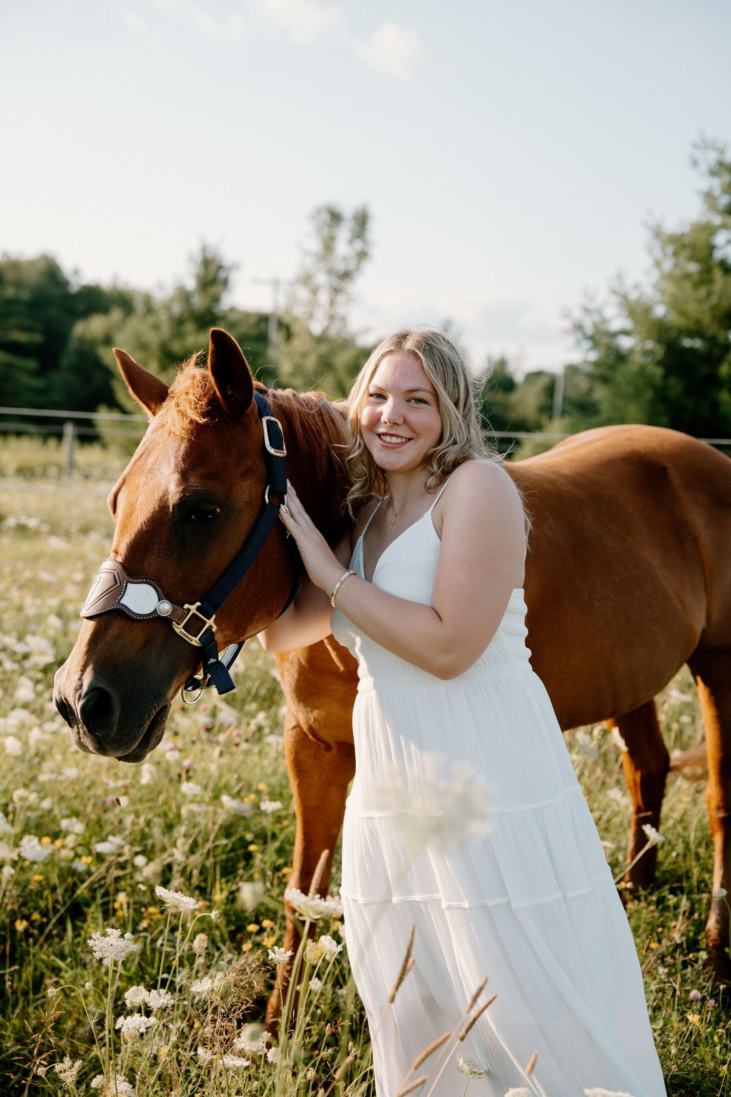 Smiling blonde woman in white dress posing with chestnut horse in sunny wildflower field.