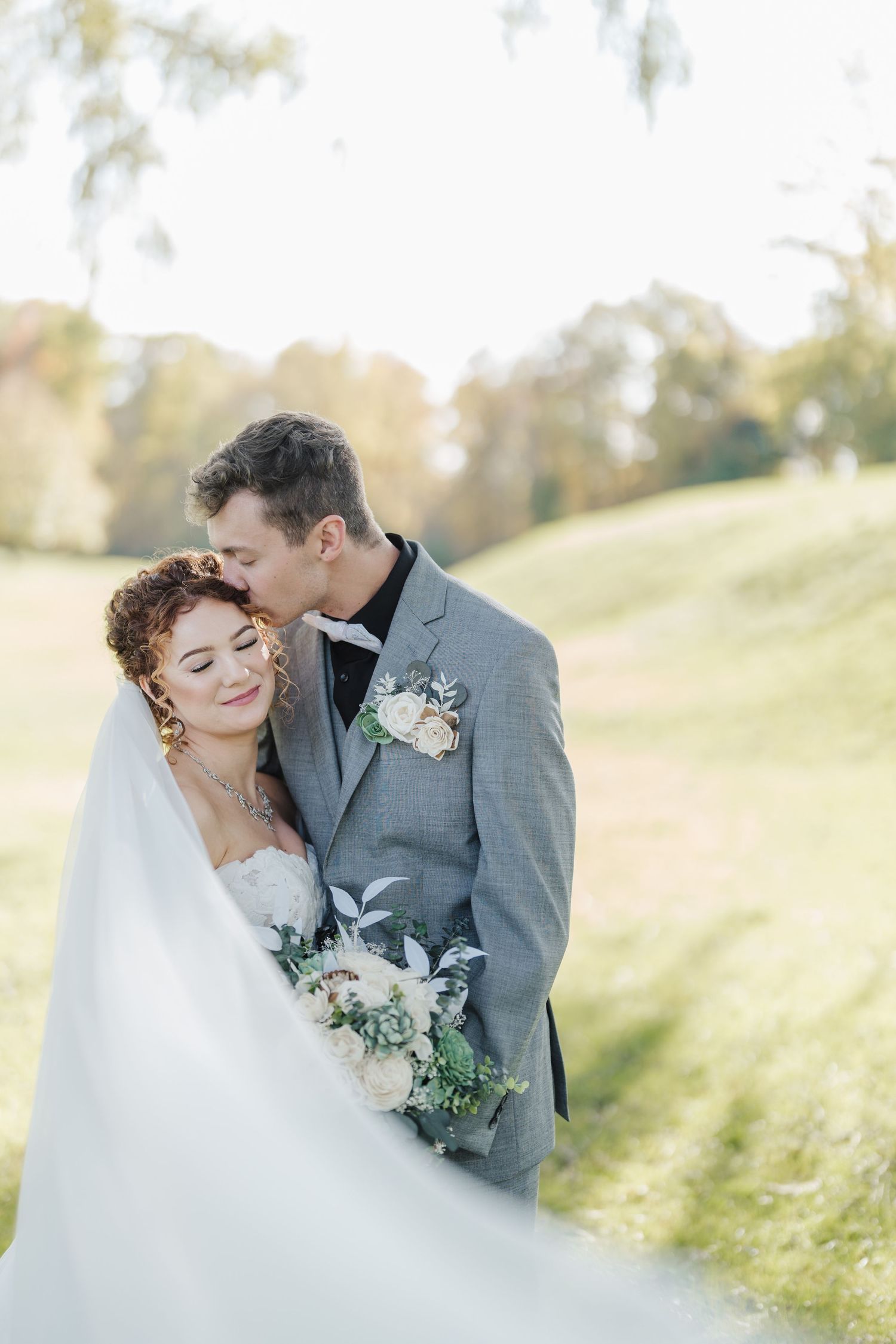 A romantic wedding photo of newlyweds sharing an intimate moment on a grassy hill in soft sunlight.