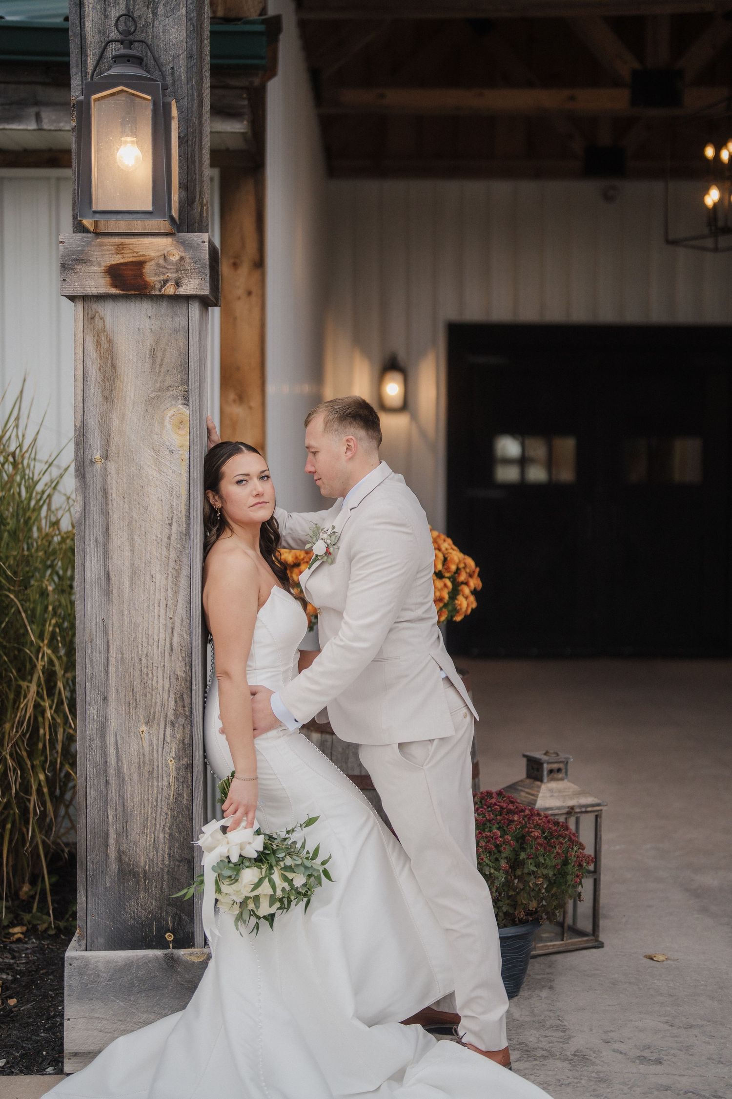 Wedding couple poses in front of rustic wooden posts with autumn decorations and string lights.