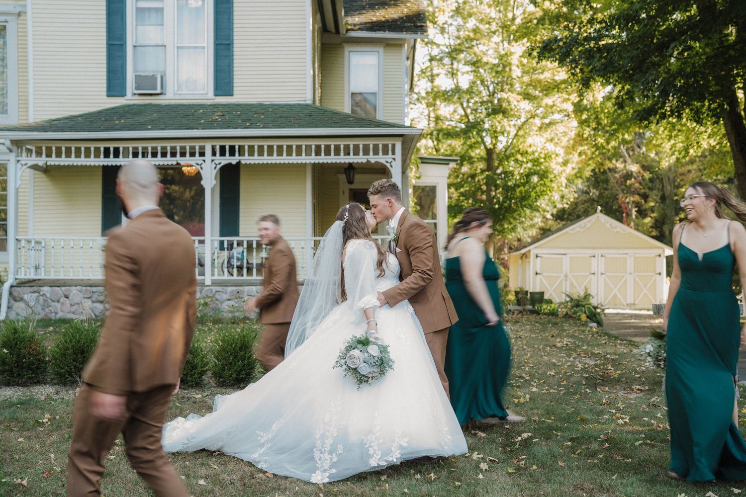 Wedding party members in teal dresses and suits pose together on the steps of a white house in golden sunlight.