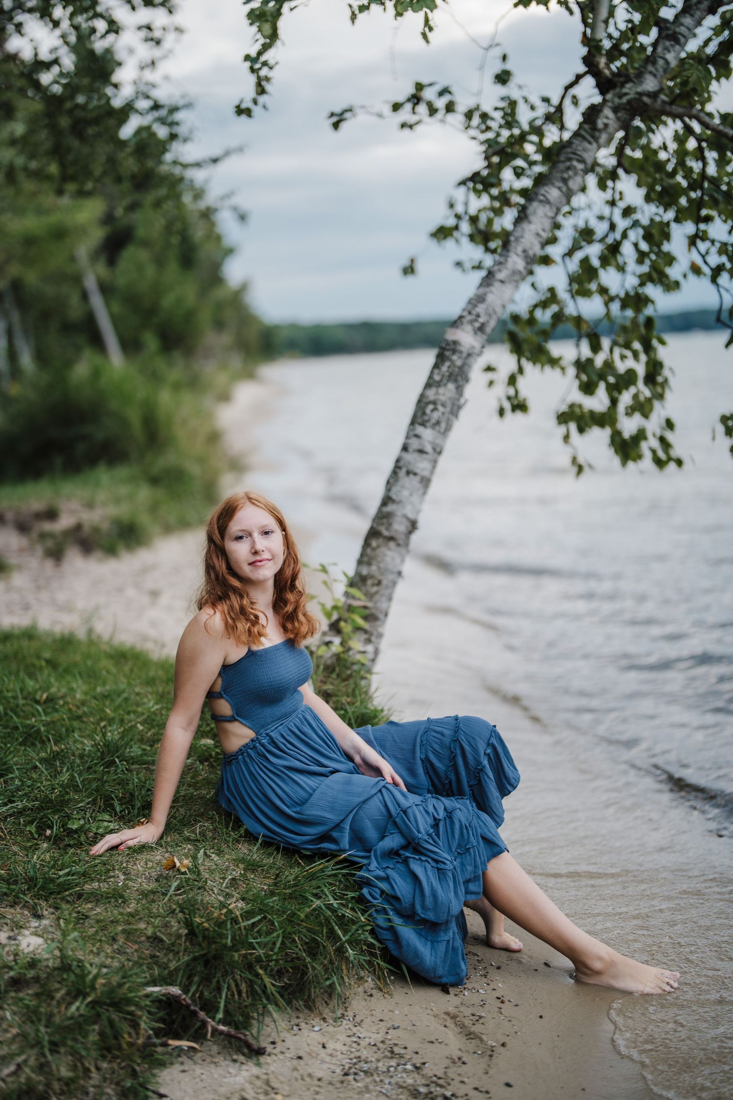 A dreamy lakeside portrait features someone reclining on a grassy shore in an elegant navy blue dress.