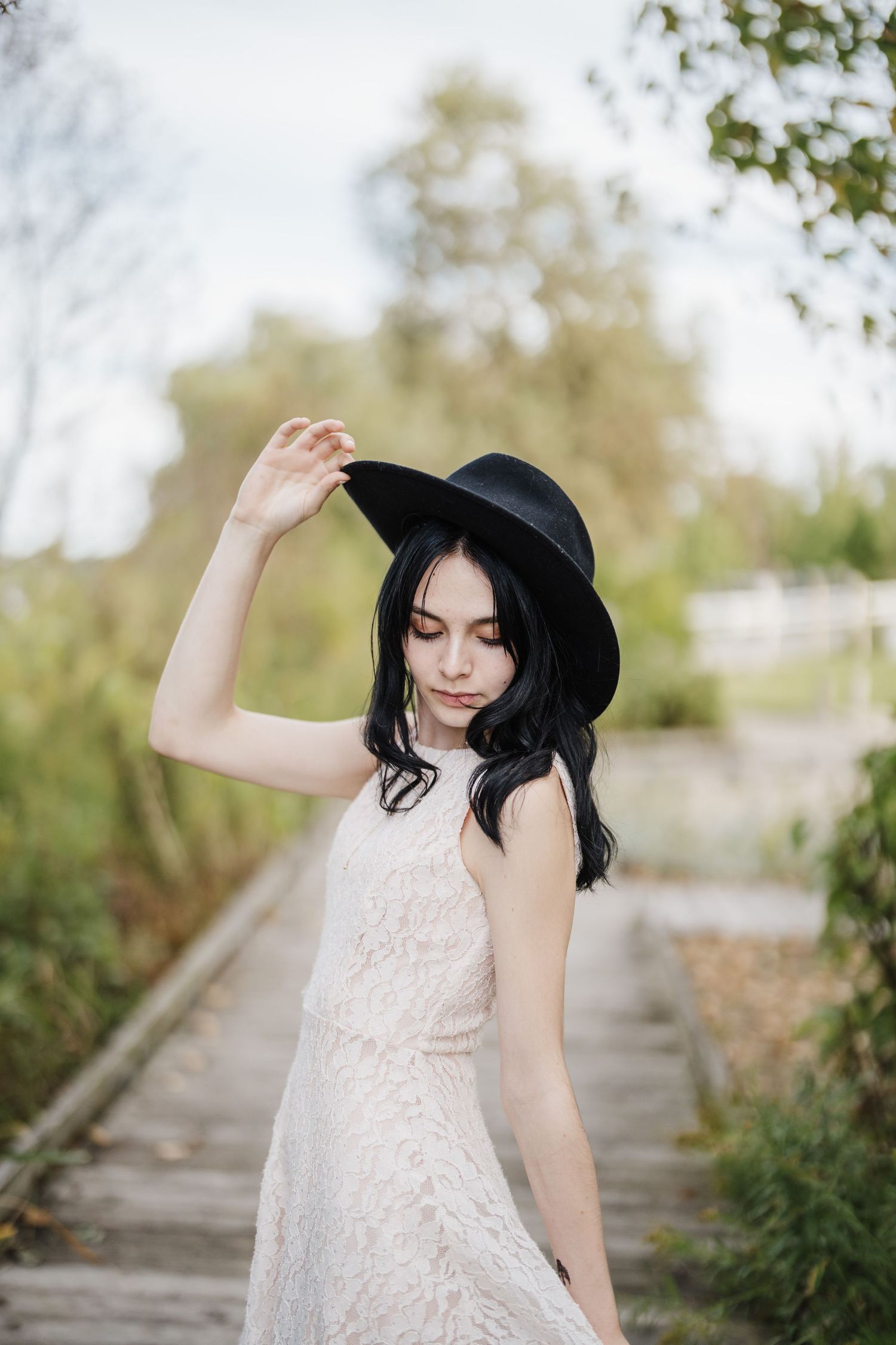 A person in a white dress and black hat twirls playfully on a path surrounded by greenery.