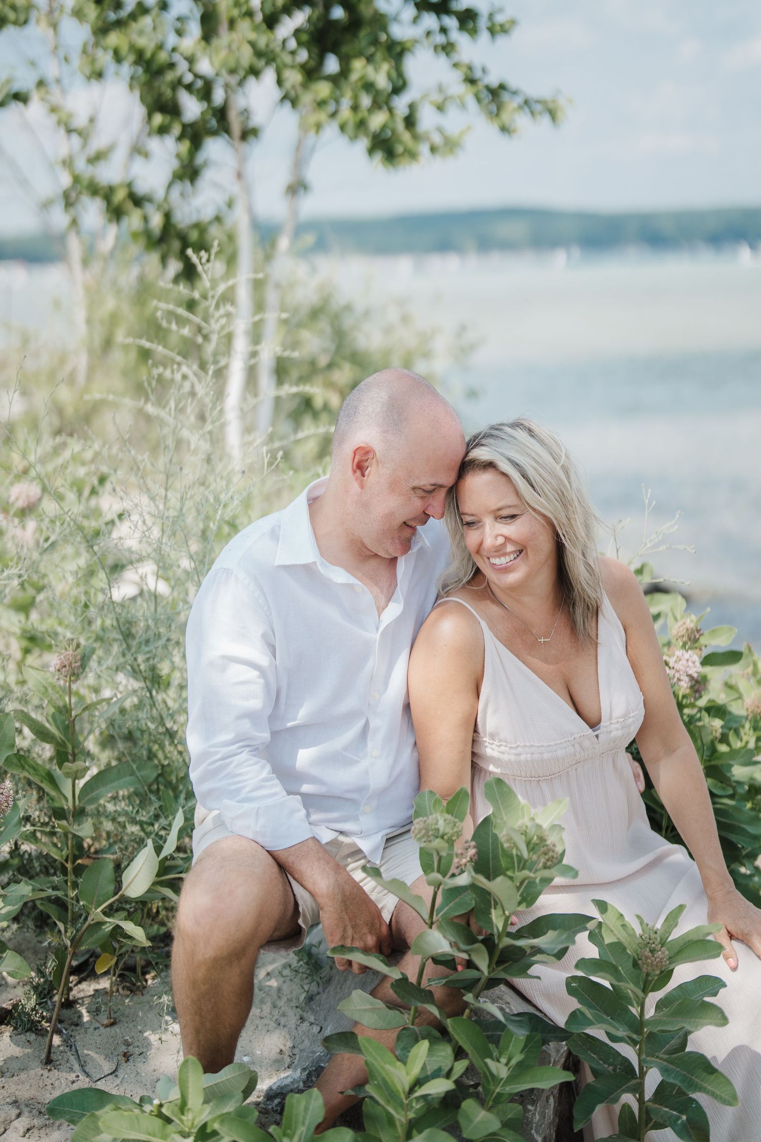 A romantic couple shares intimate moments while sitting among green foliage near a serene waterfront.