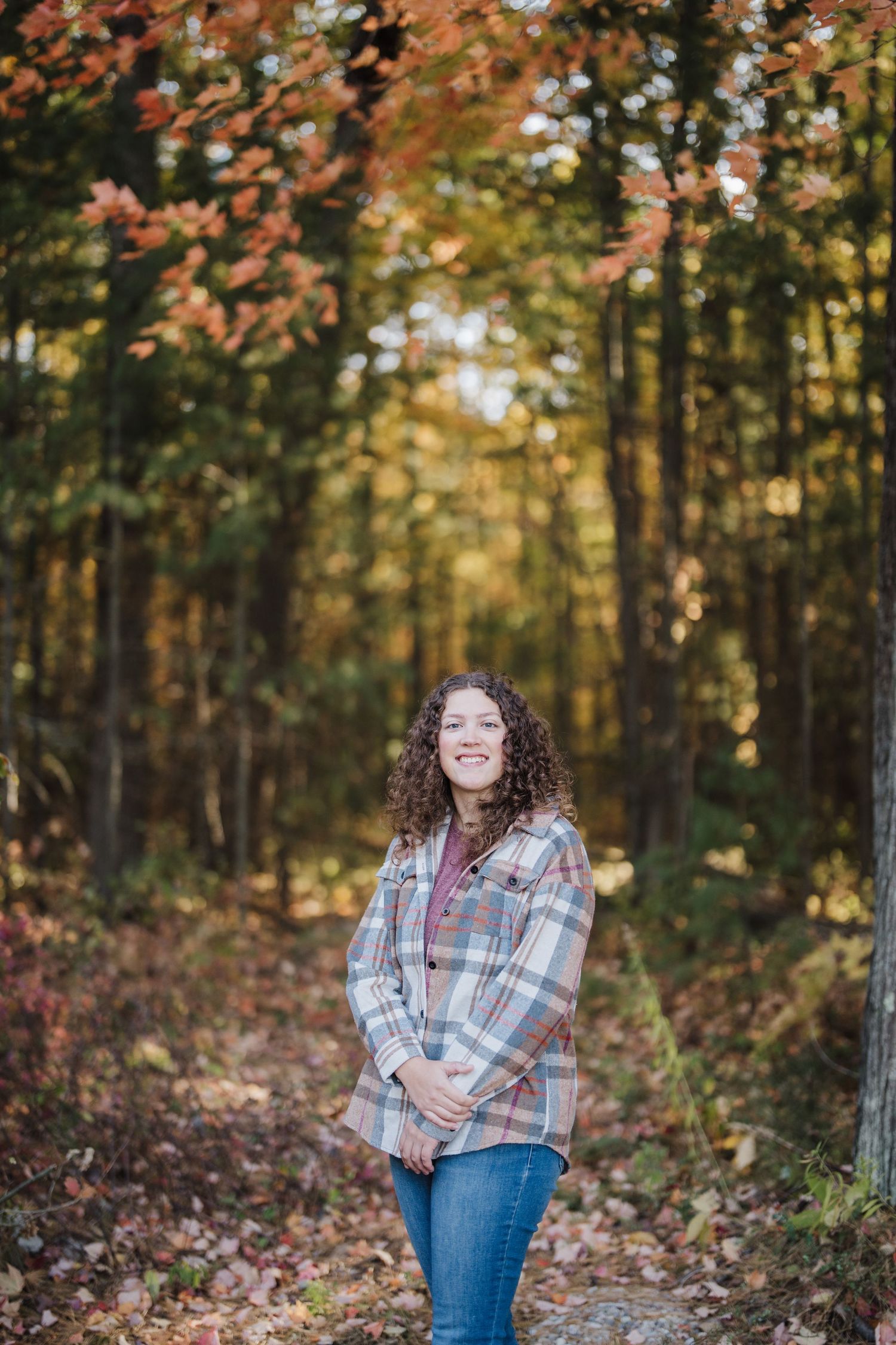 Person in plaid shirt and jeans poses in autumn forest with golden sunlight filtering through trees.