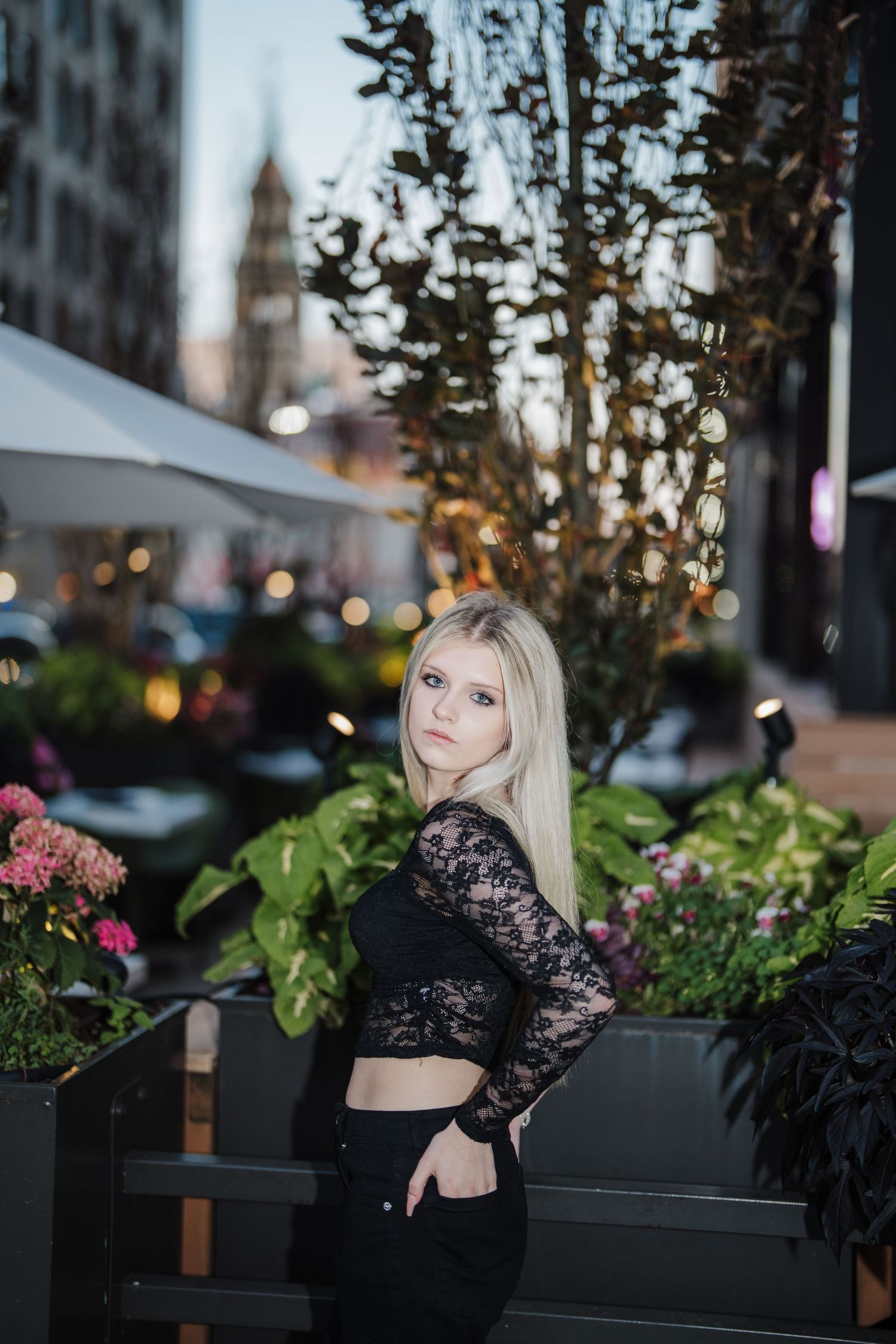 A person poses near outdoor planters with pink flowers at dusk in an urban setting.