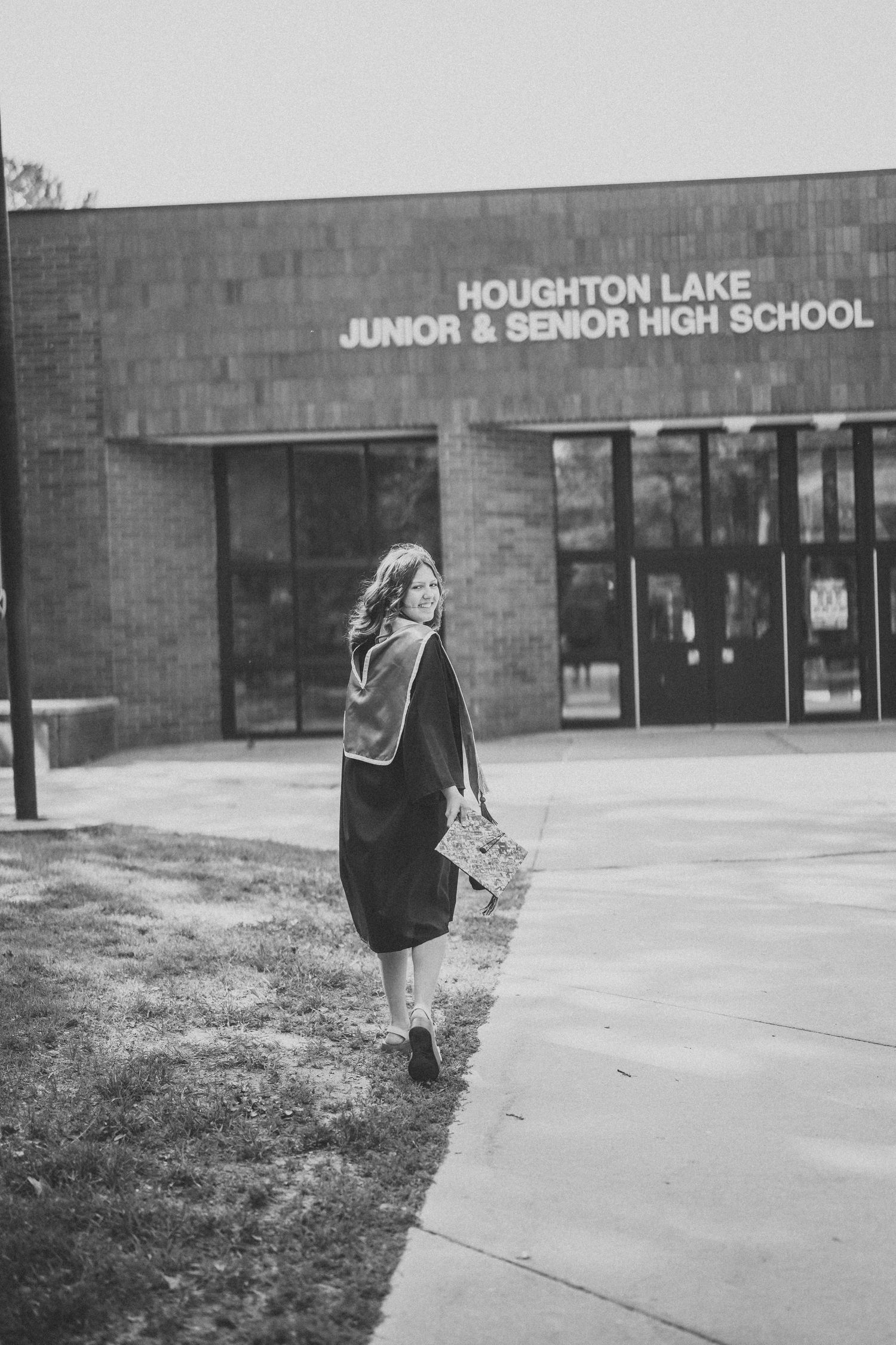 A student with a backpack walks along the sidewalk towards Houghton Lake Senior High School entrance on a cloudy day.