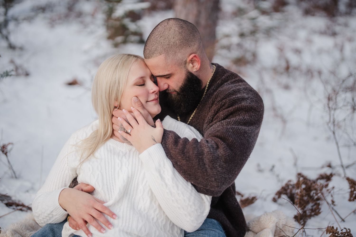 A romantic winter engagement photoshoot shows a couple embracing in the snow-covered woods.