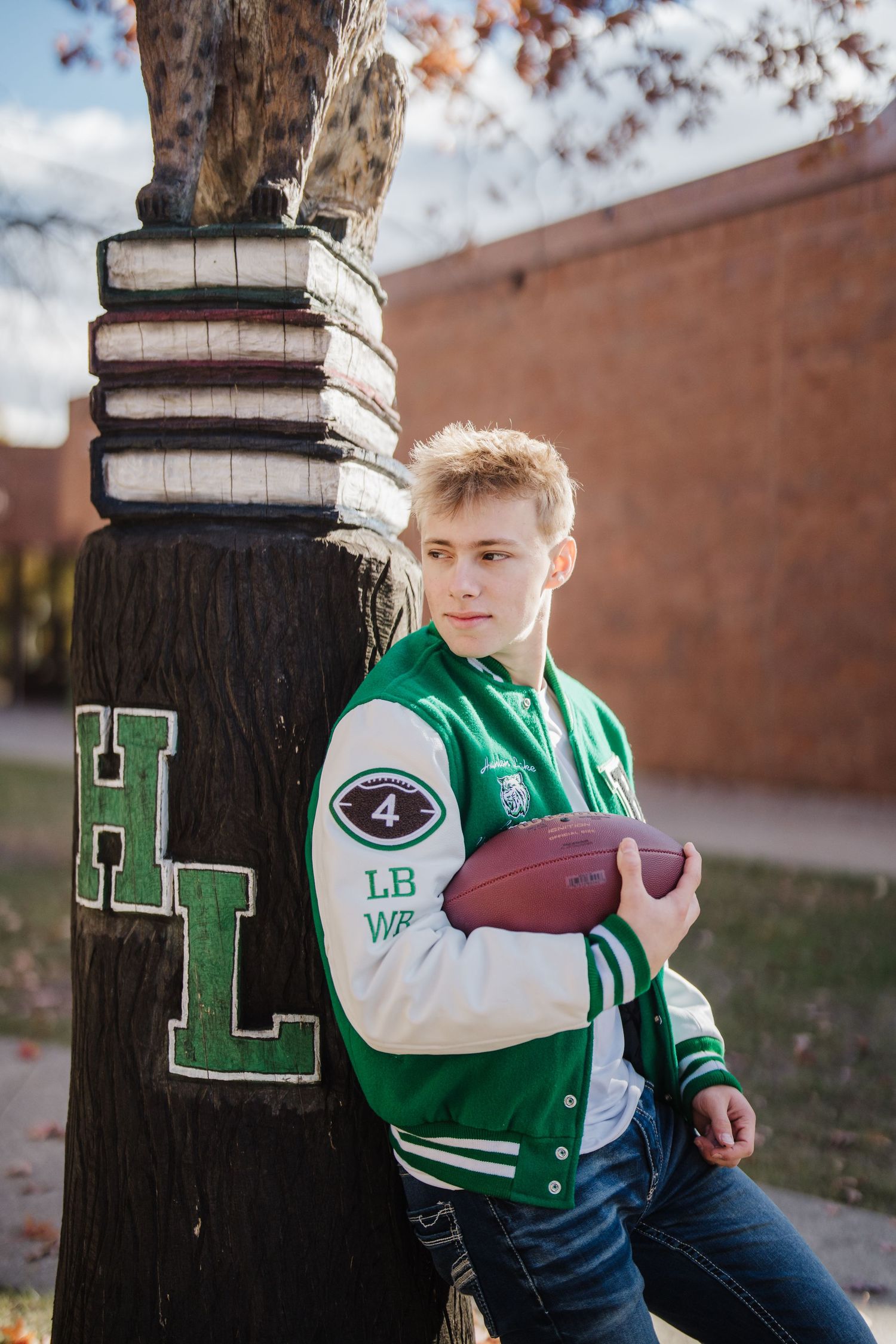 Athlete in green varsity jacket poses with football against brick wall and wooden post.