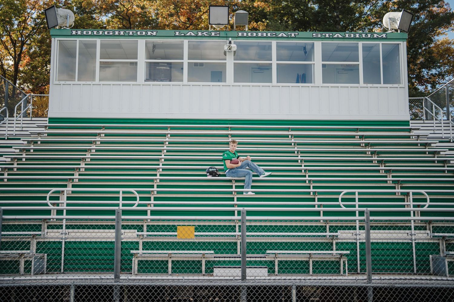 Empty green bleachers at an outdoor sports stadium with autumn trees visible in the background.