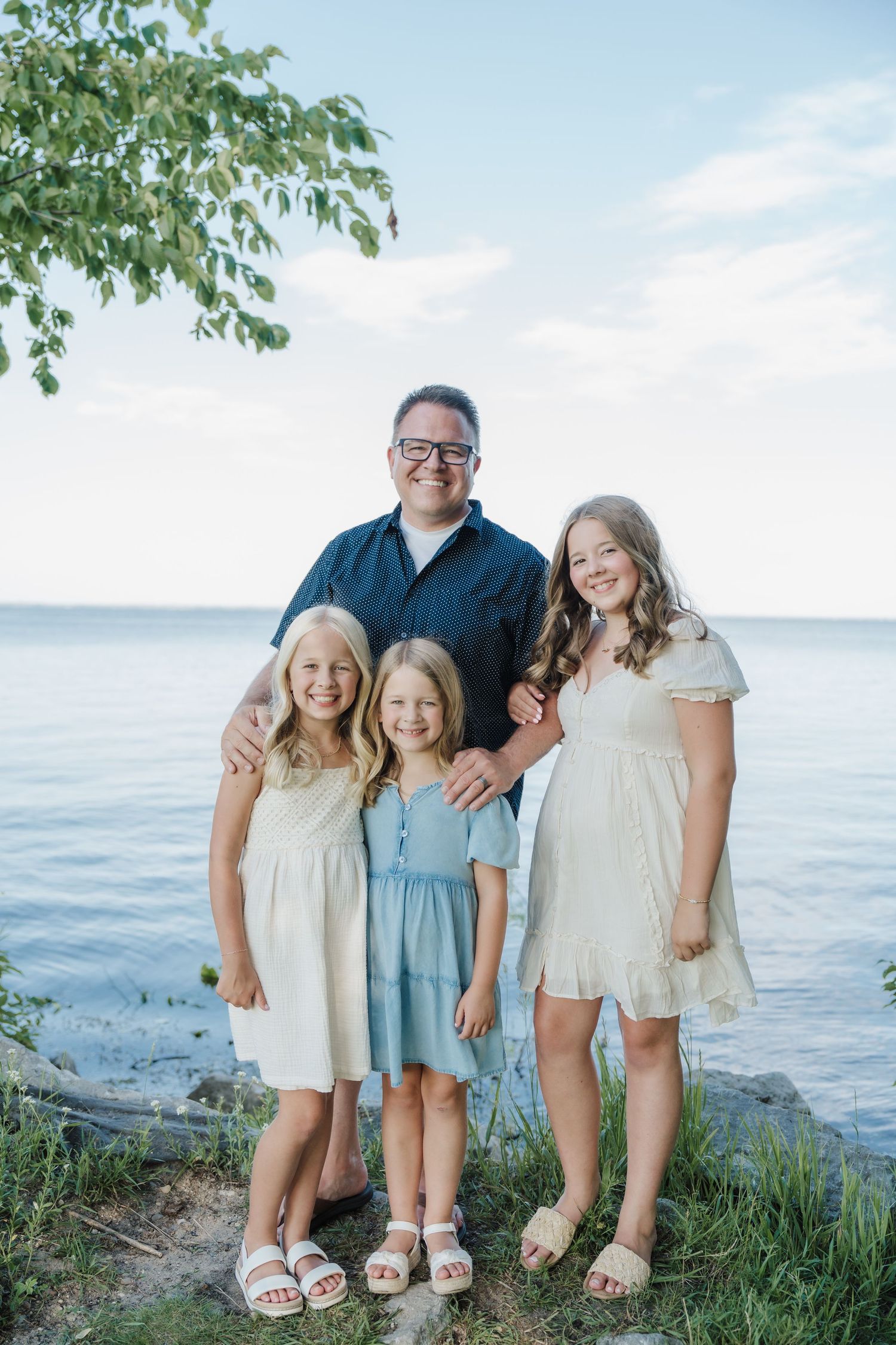 A man and three girls pose smiling by a lakeside, wearing casual summer outfits in a warm family portrait.