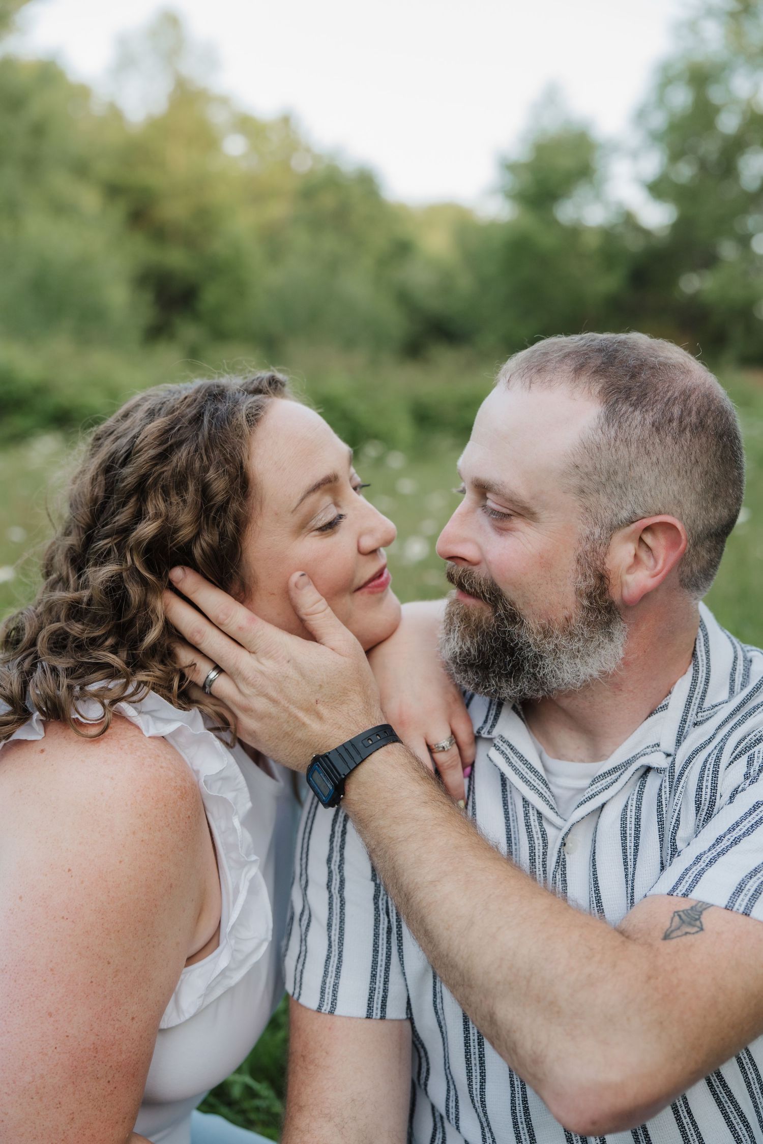 A bearded man tenderly cups a curly-haired woman's face as they gaze at each other in a lush green outdoor setting.