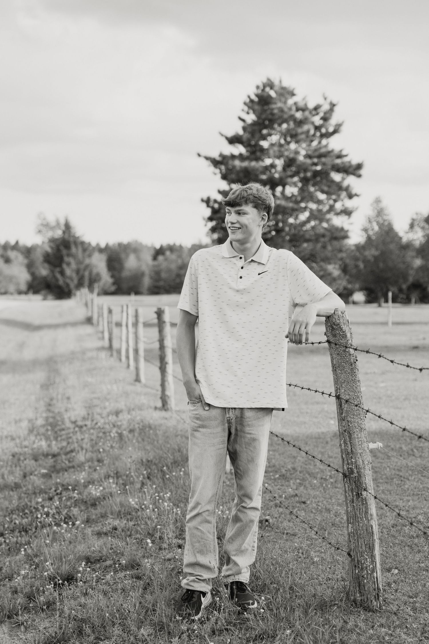 Black and white photo of a teenage boy leaning on a fence post in a rural field, smiling and wearing a polo shirt.
