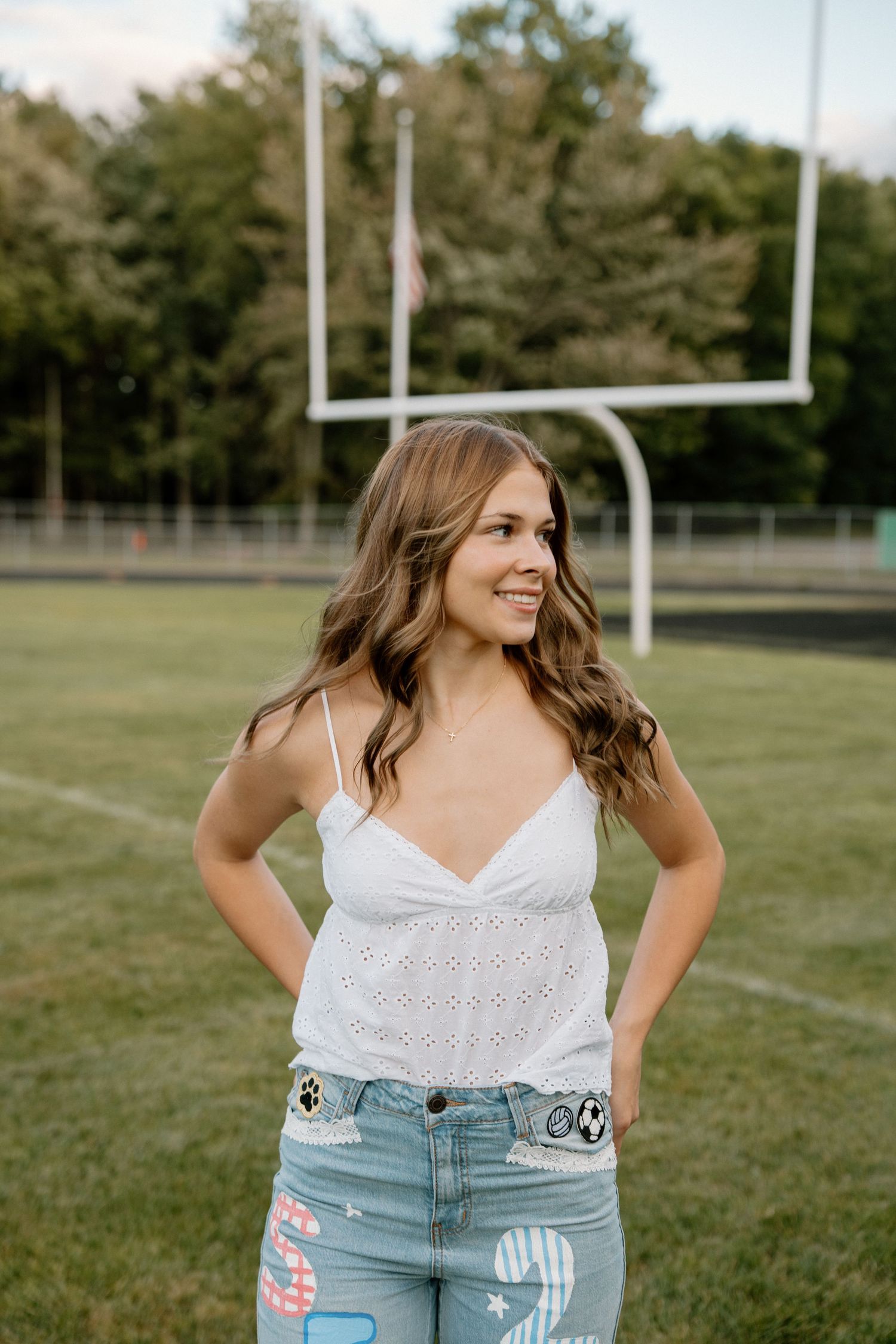 Young woman in white eyelet top and painted jeans smiling on a football field with goalposts behind her.