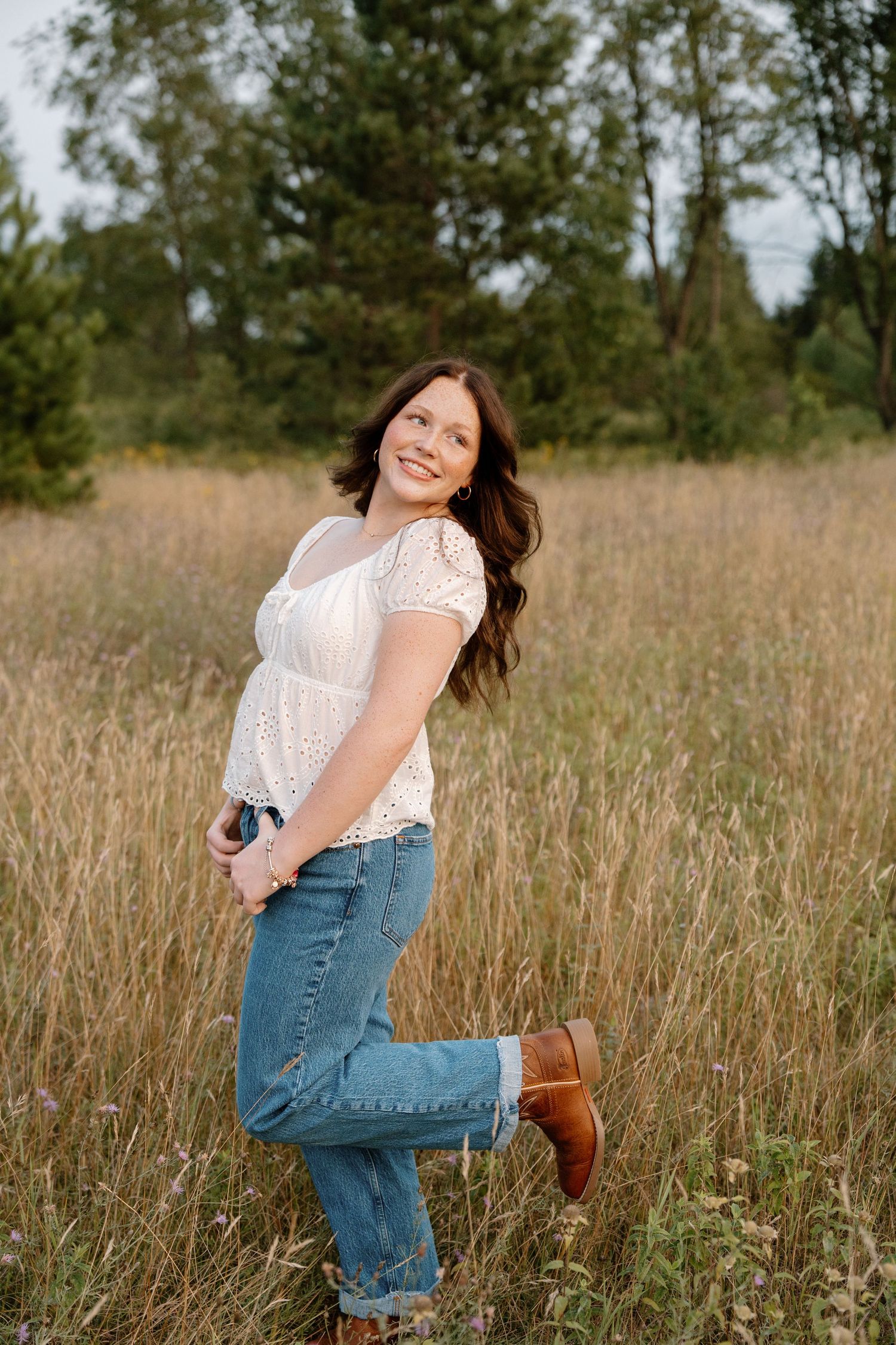 Smiling brunette woman in white top and jeans posing in a sunlit meadow.