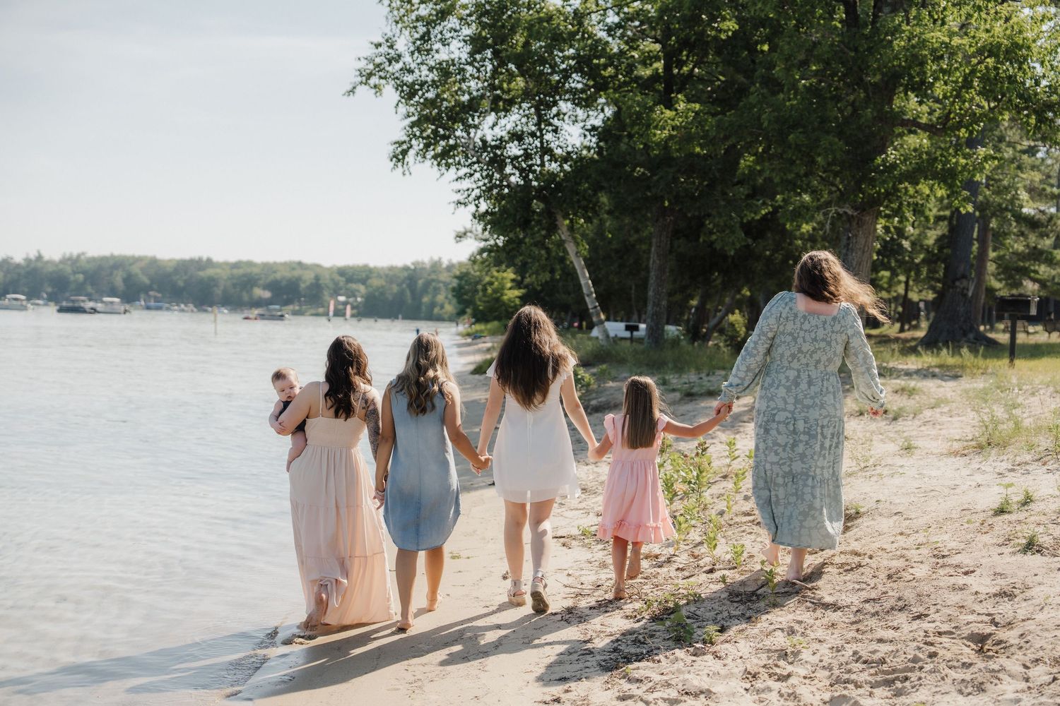 Five women and girls holding hands walk along a sandy lakeside beach surrounded by lush green trees.