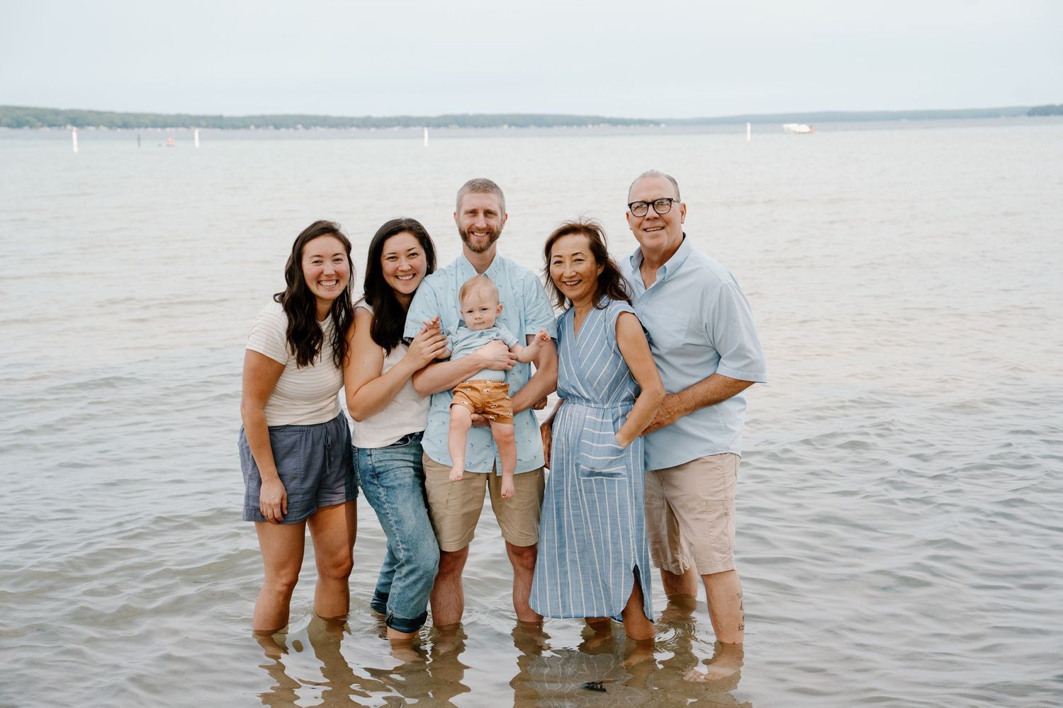 Multi-generational family smiling together in shallow ocean water at beach, holding newborn baby on cloudy day.