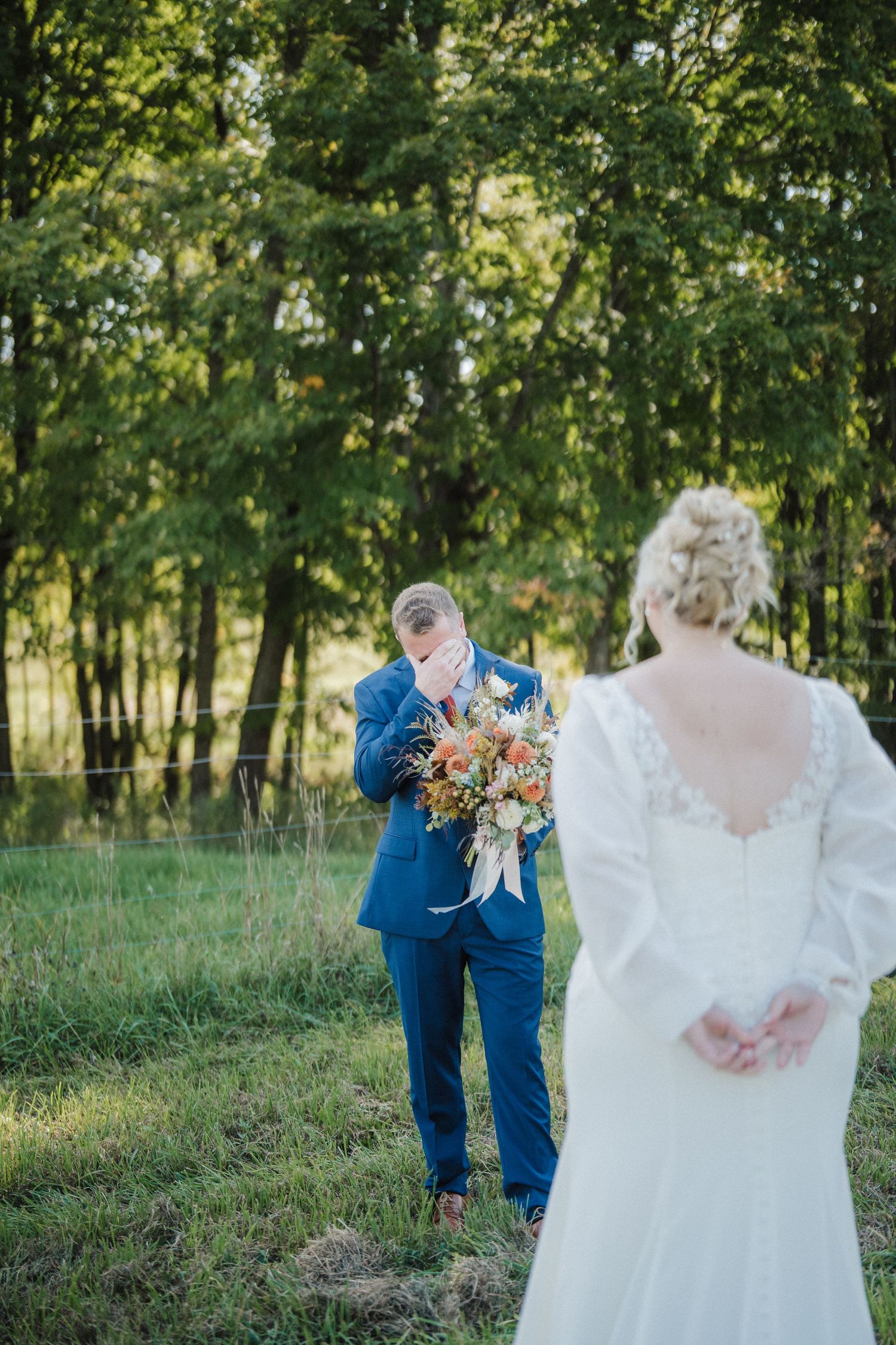 A couple in wedding attire walks through a sunlit outdoor setting with trees and grass in the background.