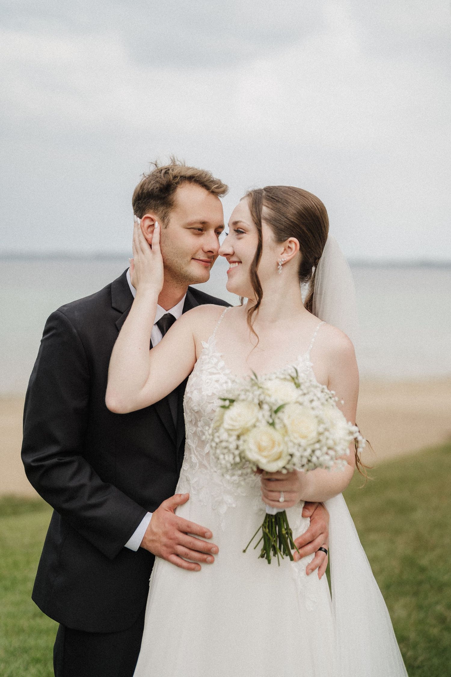 A romantic wedding couple embraces outdoors while holding a white floral bouquet on a cloudy day by the water.