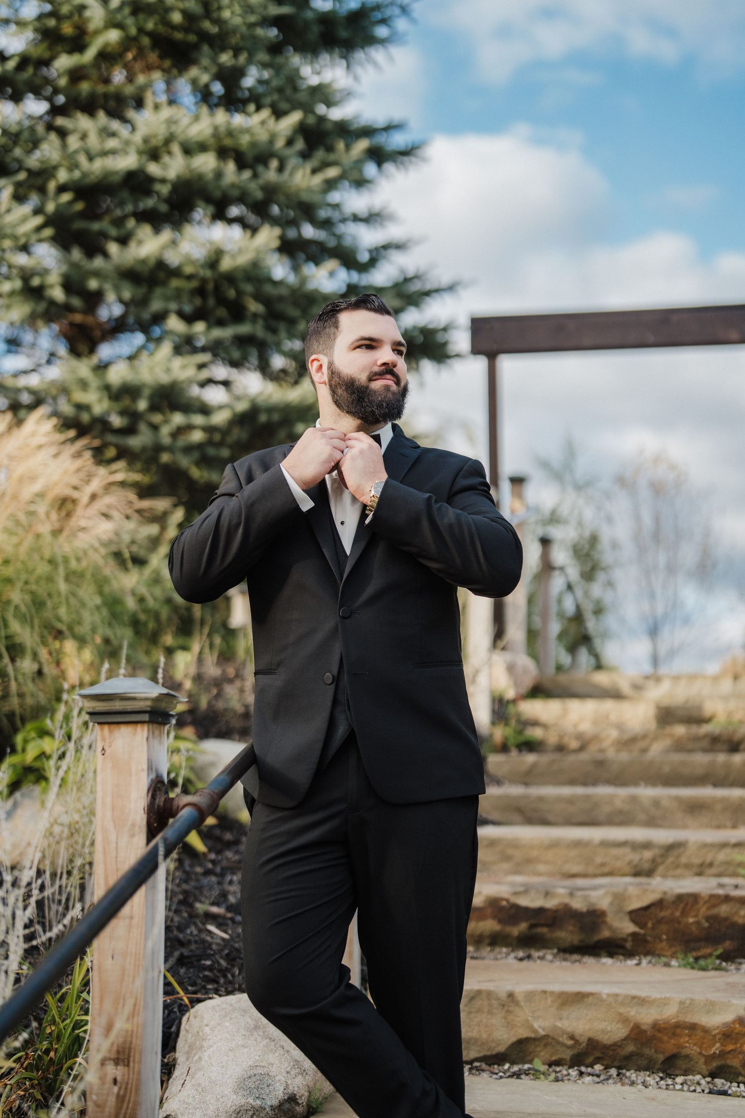 Groom in black tuxedo stands alone in an outdoor garden setting with pine trees.
