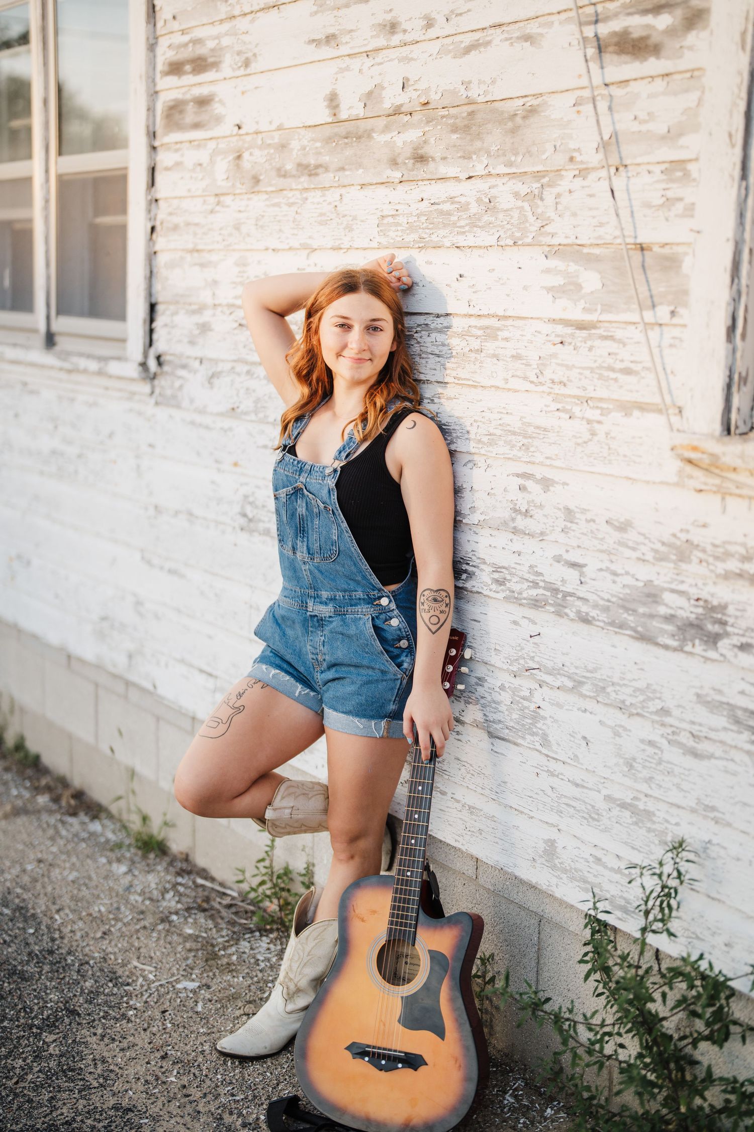 A guitarist poses with an acoustic instrument against a rustic white building exterior.