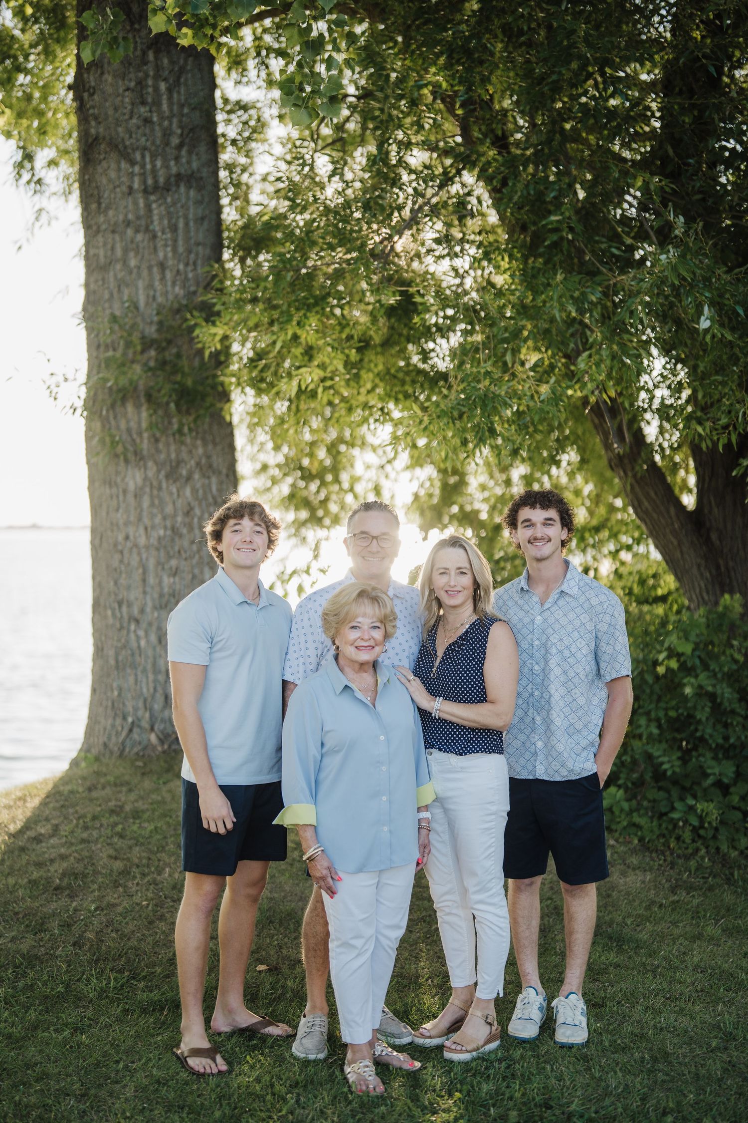 A family group poses together outdoors on a grassy area under large trees with sunlight filtering through the leaves.