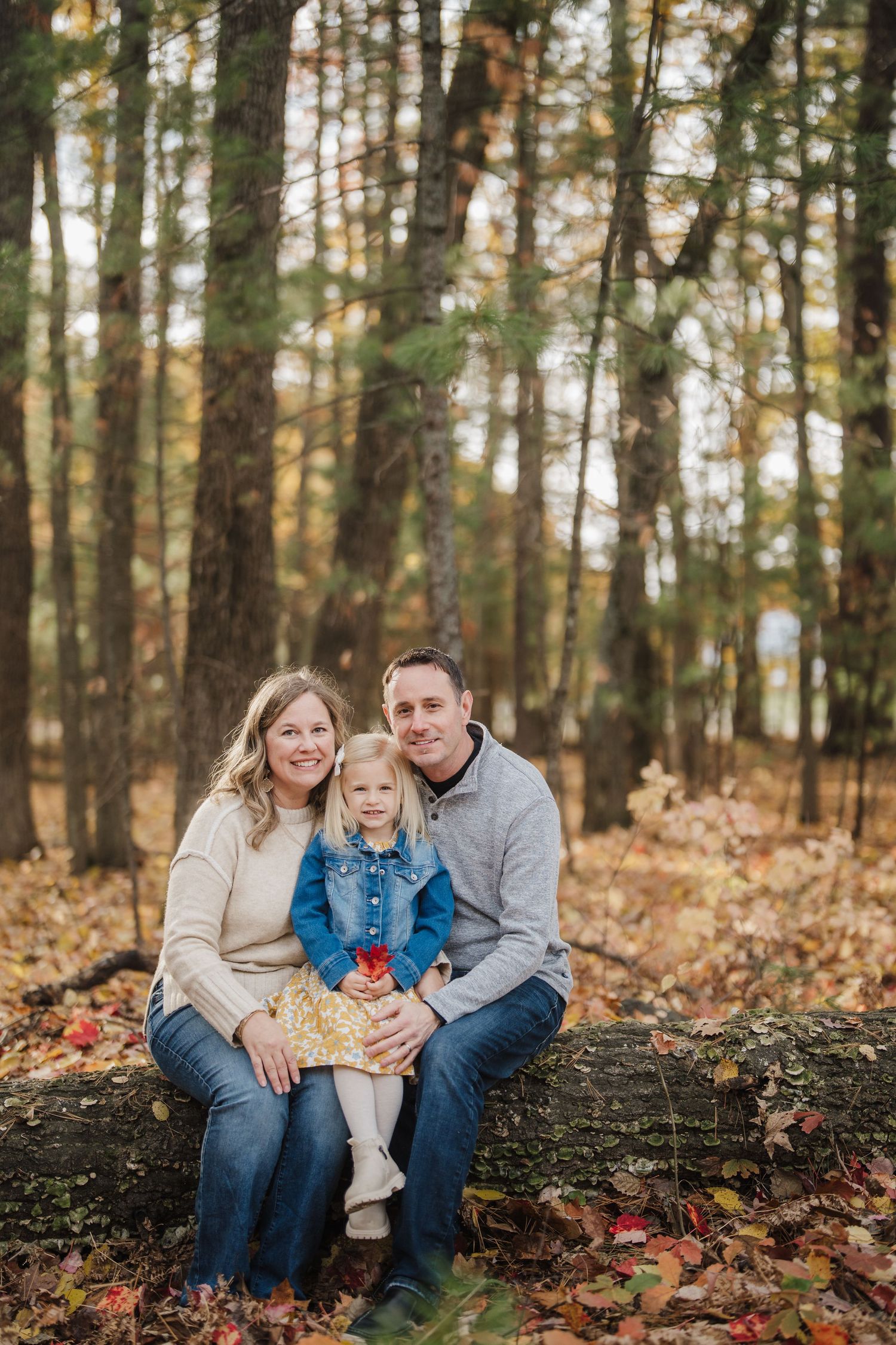 A family sits together on a fallen log in an autumn forest setting with golden leaves scattered around.