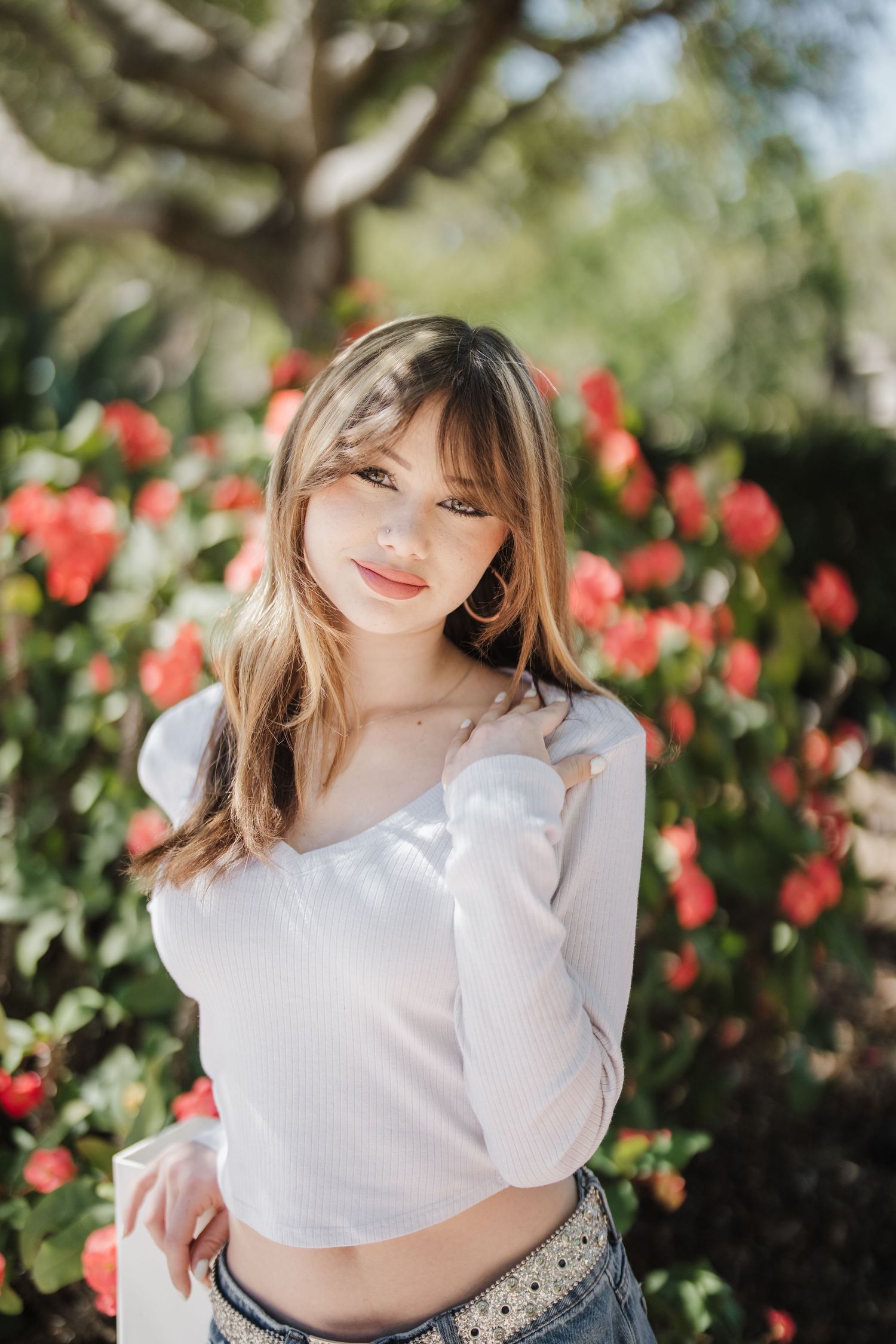 A person in a white top poses outdoors against a background of vibrant red flowers and greenery.