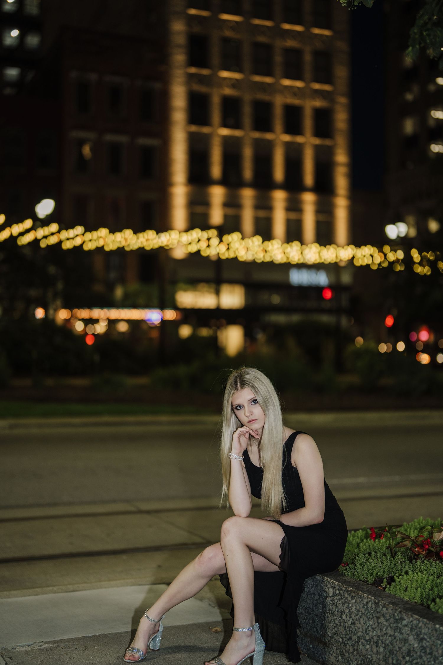 A person in a black dress sits on a stone ledge at night with illuminated buildings in the background.