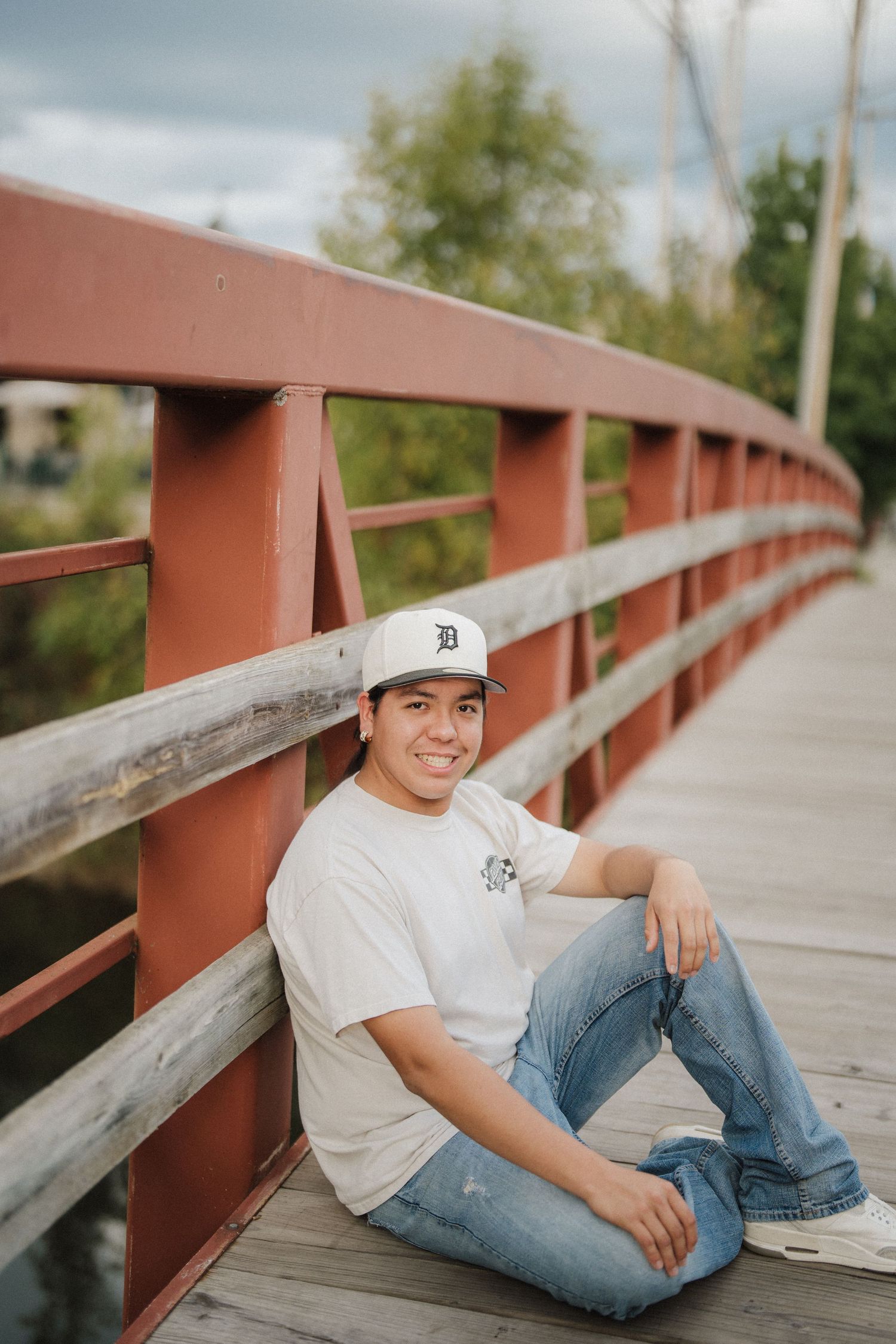 Person in Detroit Tigers baseball cap and jeans lounges on wooden bridge railing in natural outdoor setting.