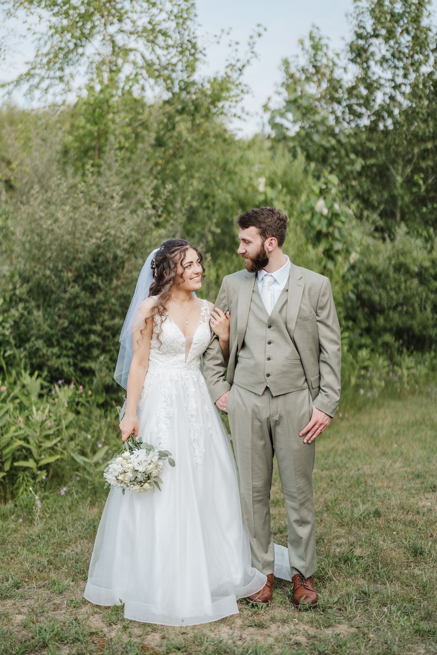 Bride and groom in garden setting, she in lace gown and he in light grey three-piece suit.