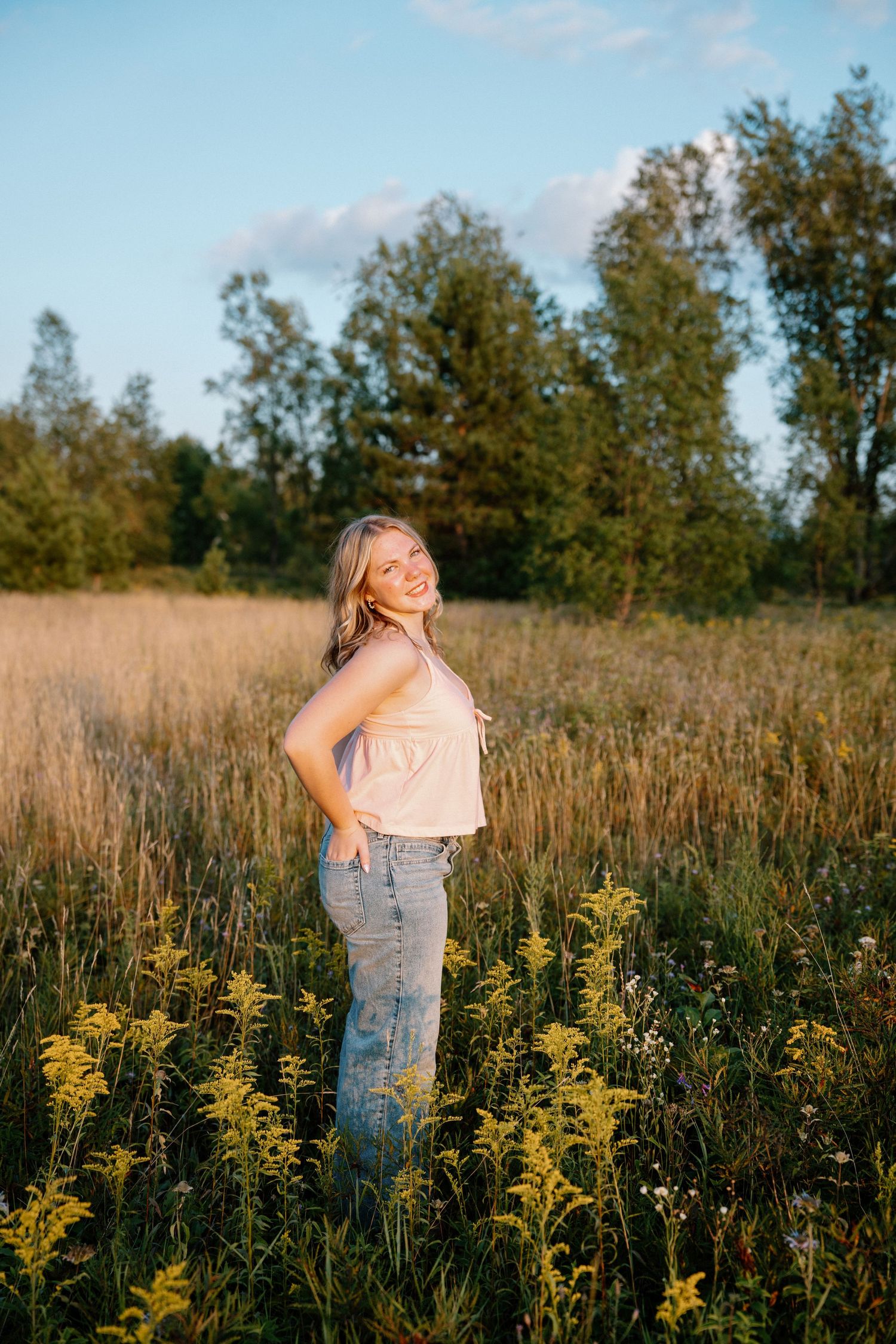 Blonde woman in pink top and jeans smiling in golden hour meadow with wildflowers.