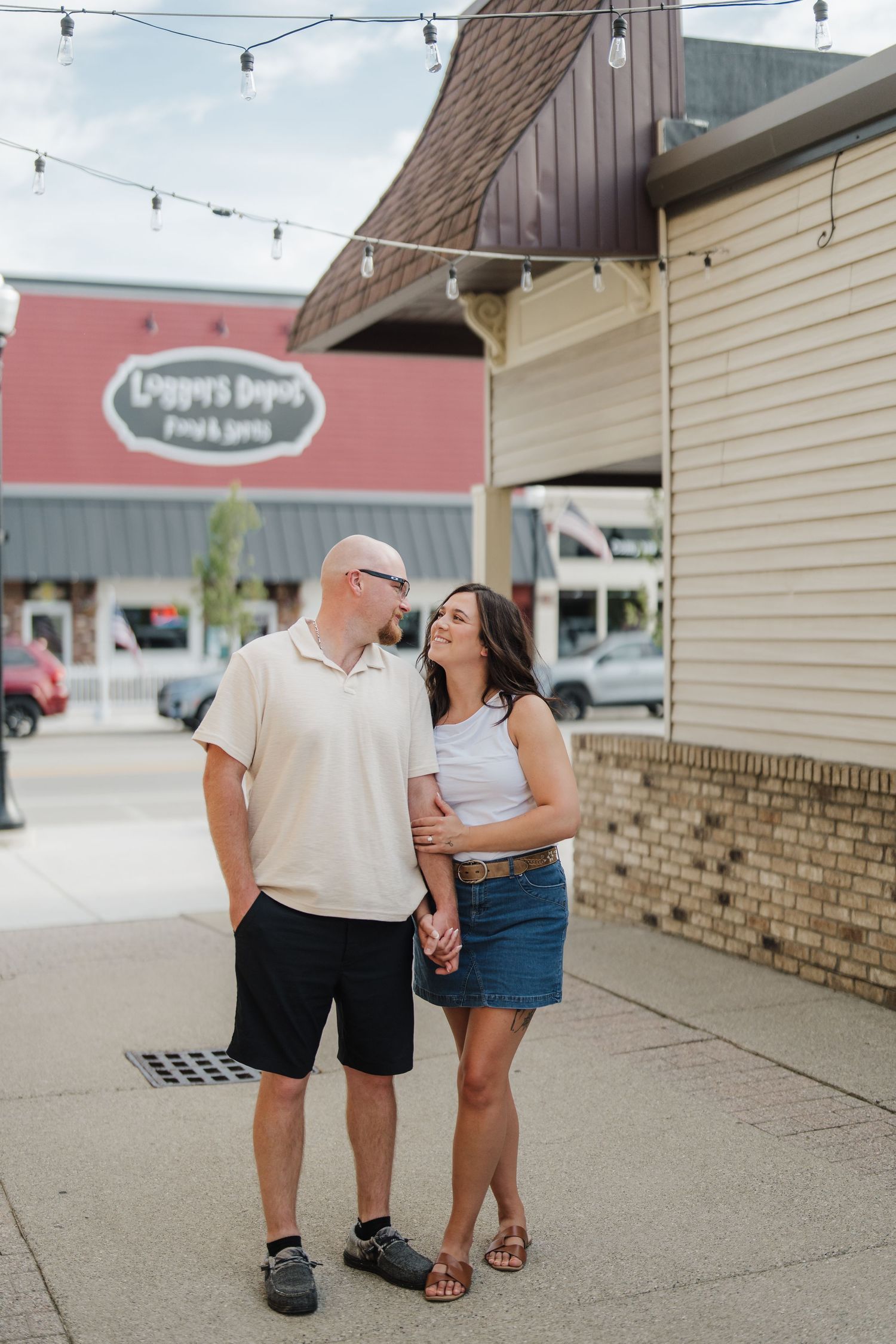 Couple holding hands and smiling at each other on a sidewalk outside a shop with string lights overhead.