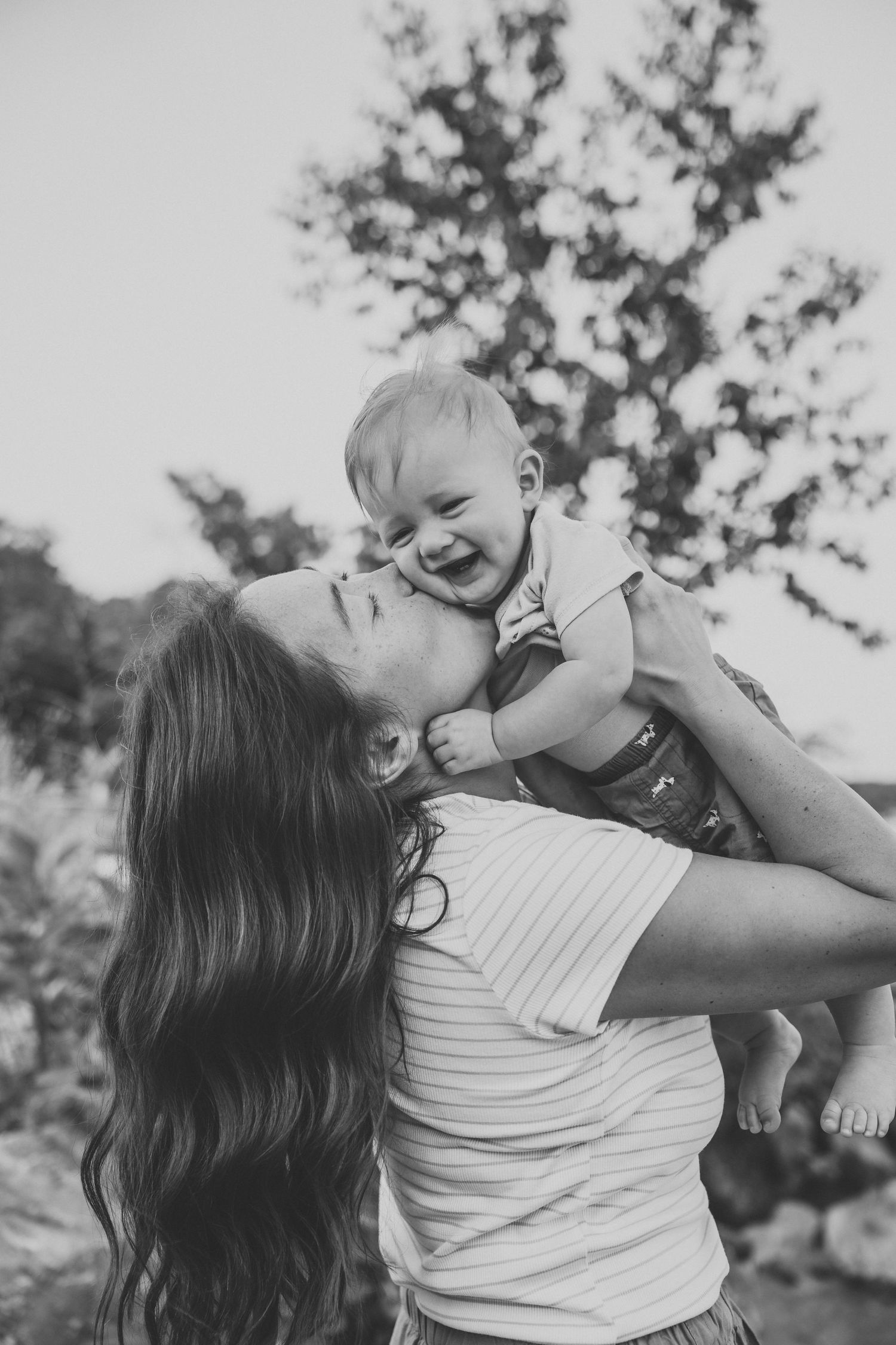 Black and white photo of a joyful mother lifting her laughing baby outdoors with trees in the background.