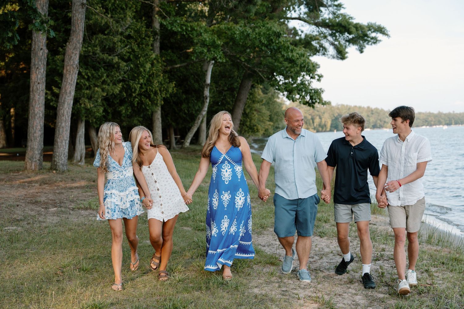 Family of six walking along a lakeside beach path surrounded by tall pine trees on a sunny summer day.