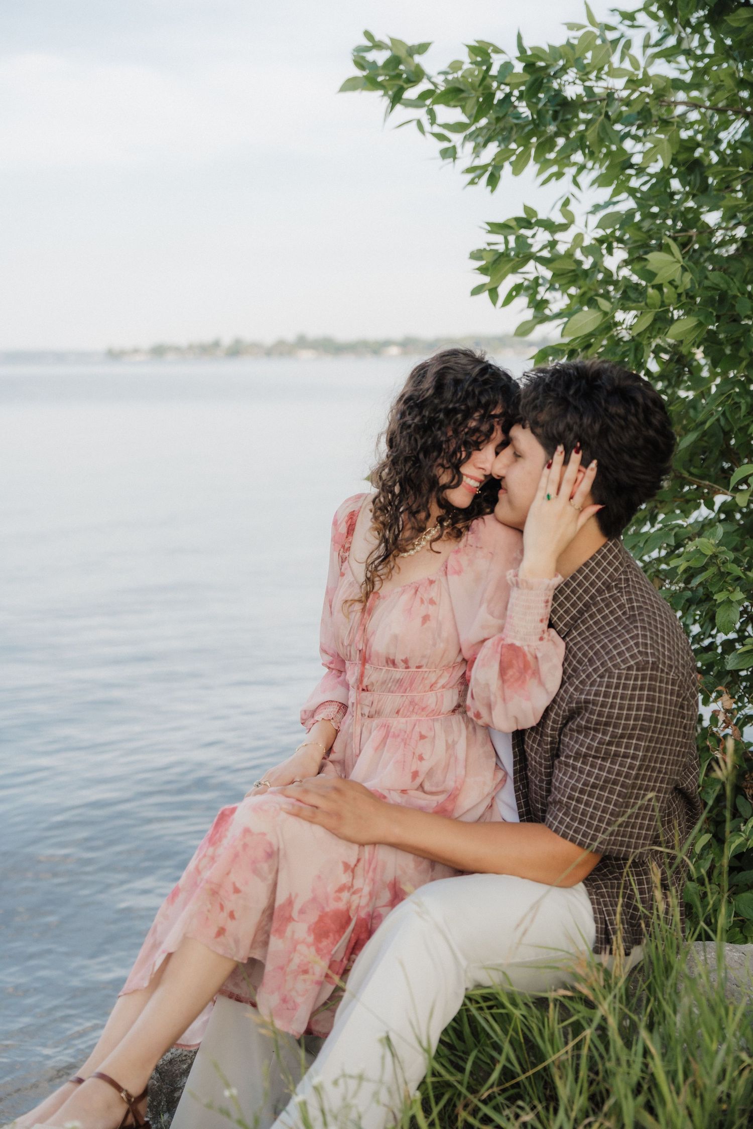 Romantic couple sharing a tender moment by the water, woman in floral pink dress, seated among green foliage.