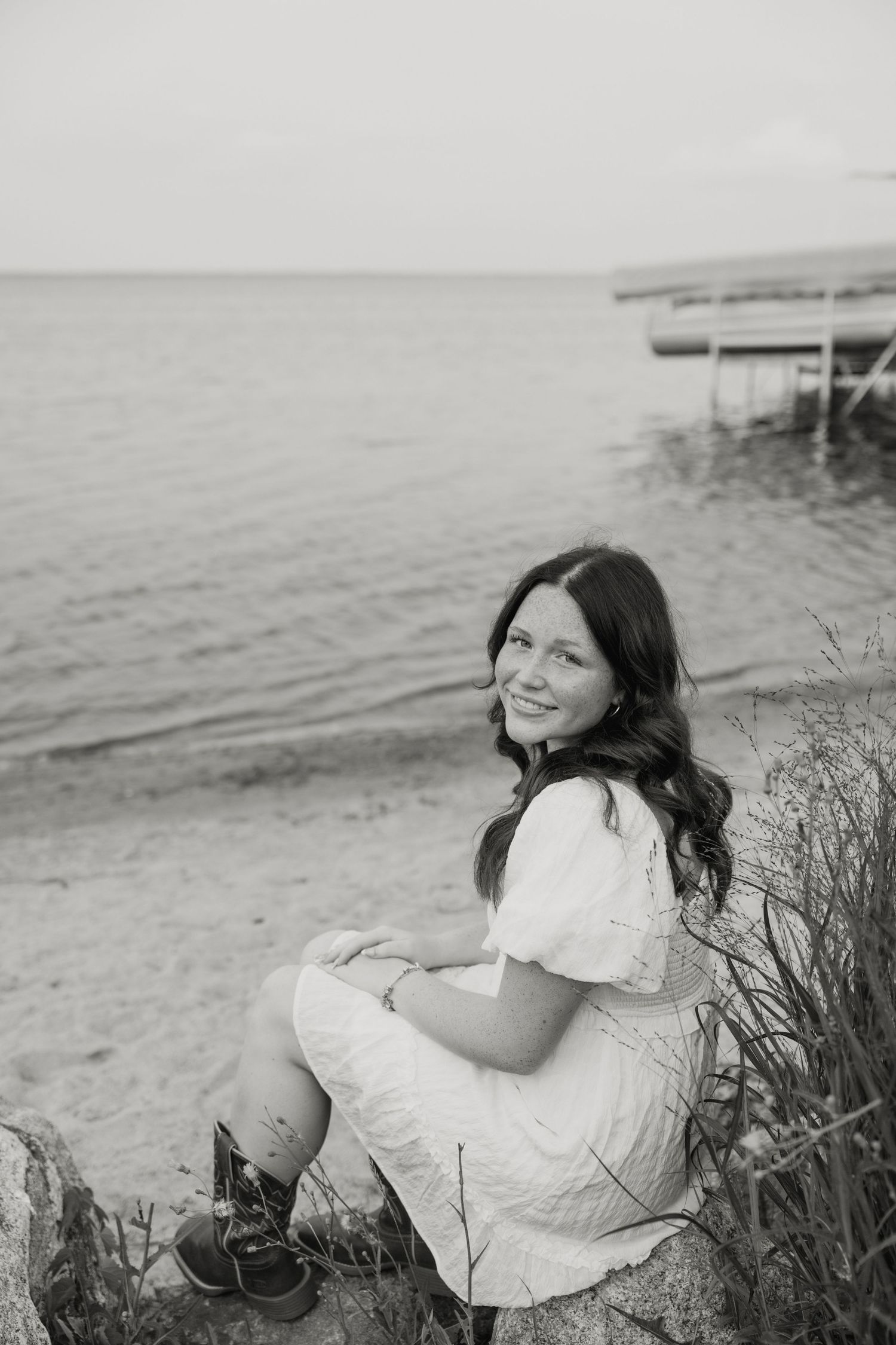 Black and white photo of a smiling young woman in a white dress sitting on rocks by a serene lake shore.